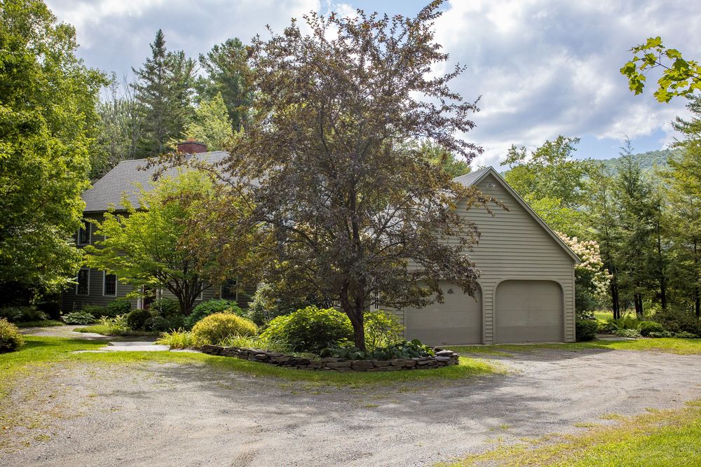 Gray New England home with attached garage surrounded by mature trees and circular gravel driveway.