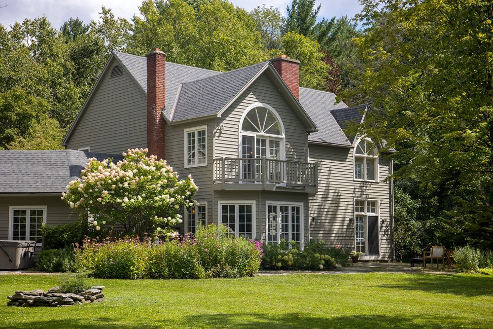 Elegant gray shingle-style home with balcony and arched window surrounded by blooming summer gardens.