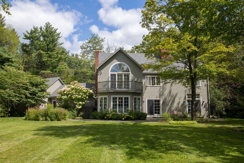 Beautiful gray shingle home with arched window, balcony, and chimney surrounded by lush mature trees.