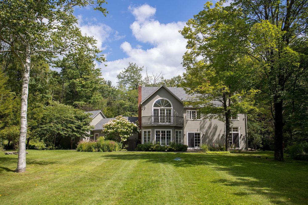 Gray two-story home featuring large arched window and balcony set among tall mature trees and lawn.