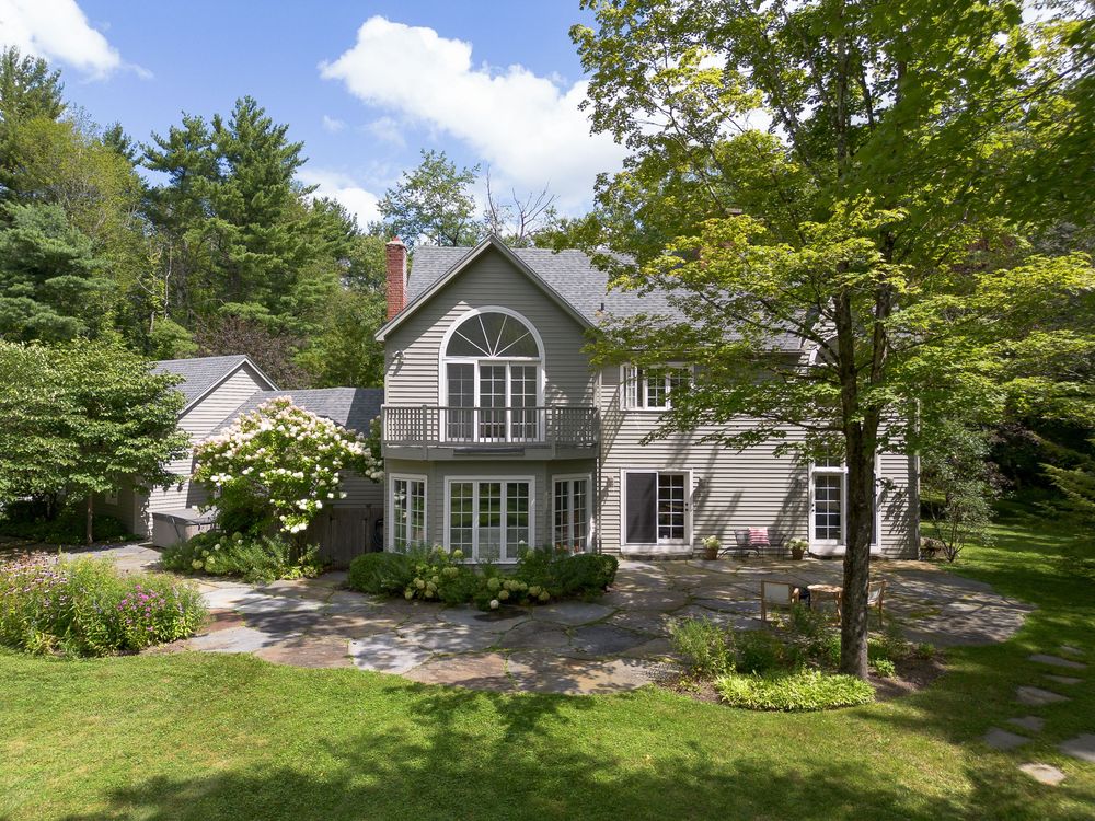 Gray colonial home with arched window, balcony, stone patio, and lush landscaping surrounded by mature trees.
