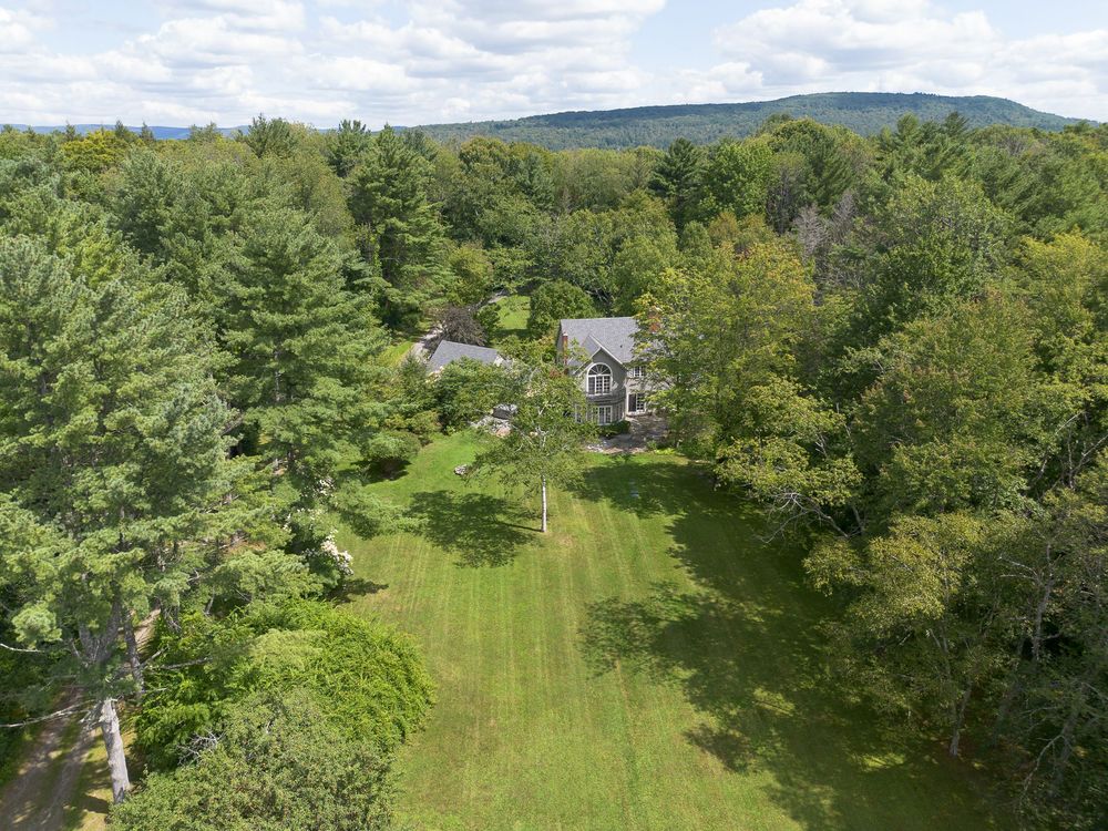 Aerial view of secluded gray colonial home nestled among dense green trees with mountain views beyond.