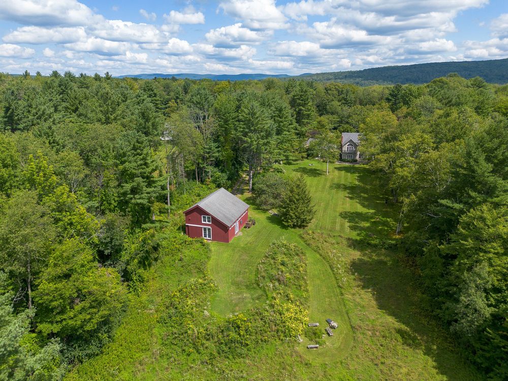 Aerial view of charming red barn surrounded by green meadows and dense forest with mountain backdrop.