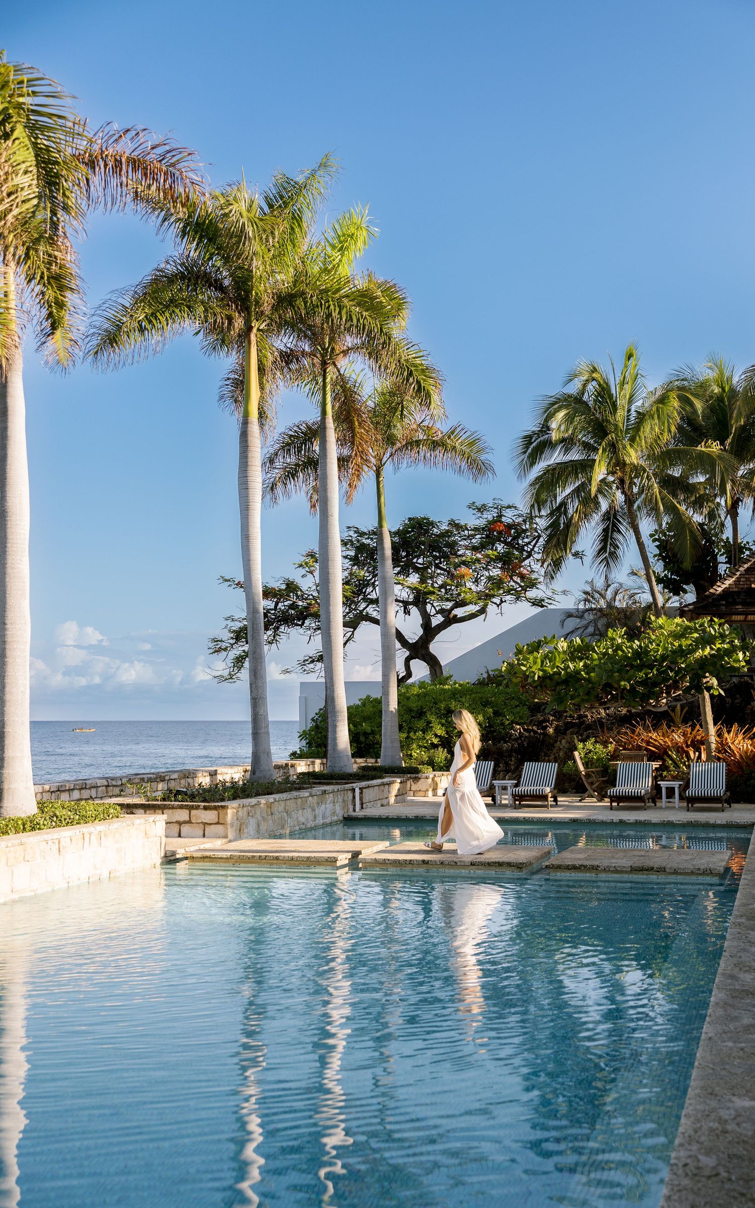 Woman in white dress walking by infinity pool with palm trees and ocean view on a sunny day.