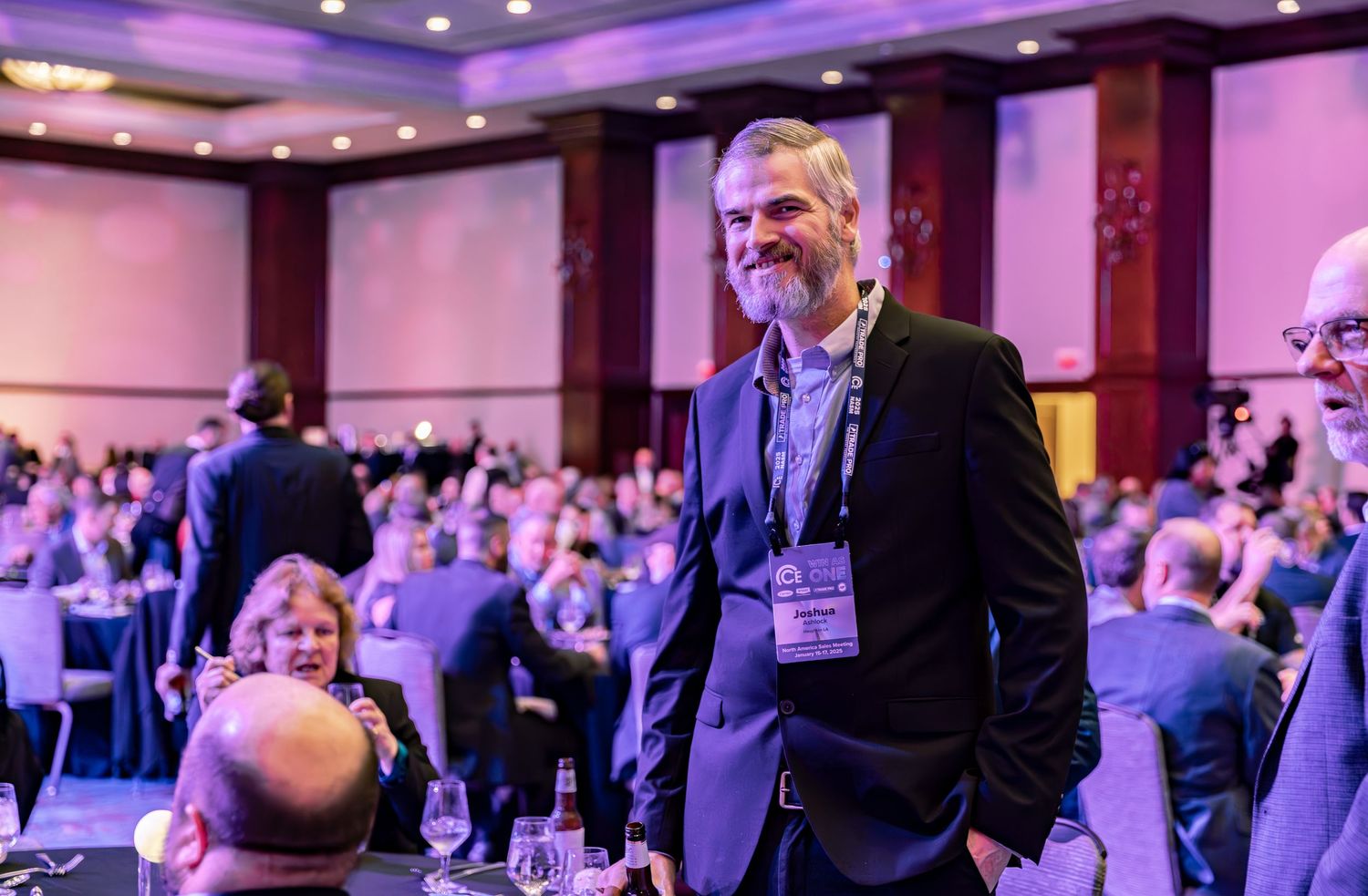 Man with beard wearing lanyard smiles at crowded conference banquet hall event.