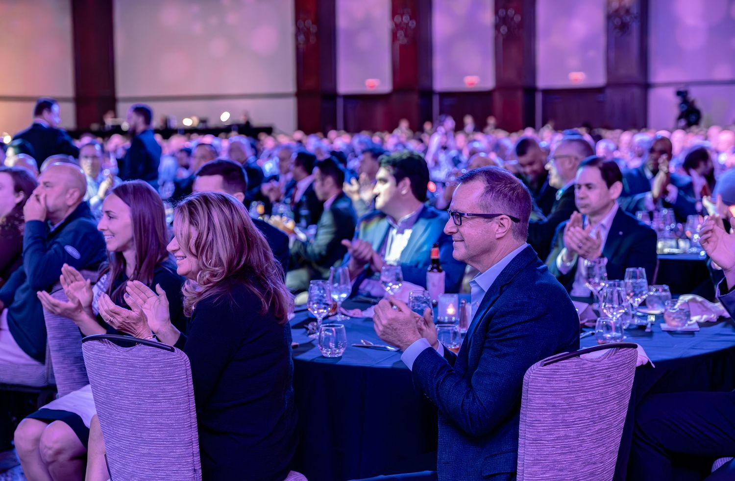 Elegantly dressed guests applaud at a formal gala dinner in a grand ballroom with purple ambient lighting.