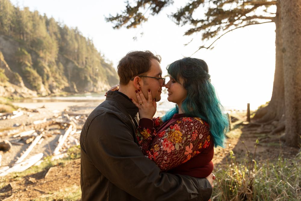 Couple sharing a romantic moment near a scenic beach with tall trees and sunlight filtering through the forest behind them.