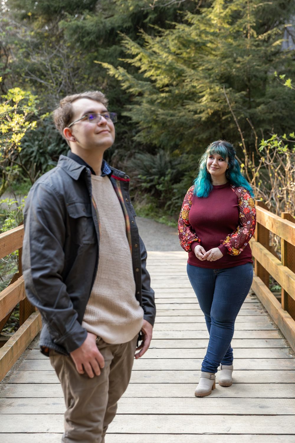 A man and woman with teal hair stand on a wooden bridge surrounded by lush green trees in a forest setting.
