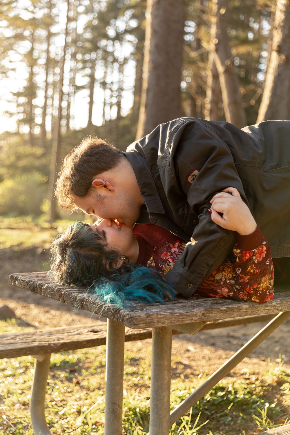 Couple sharing a romantic kiss on a rustic wooden bench in a sunlit forest setting.