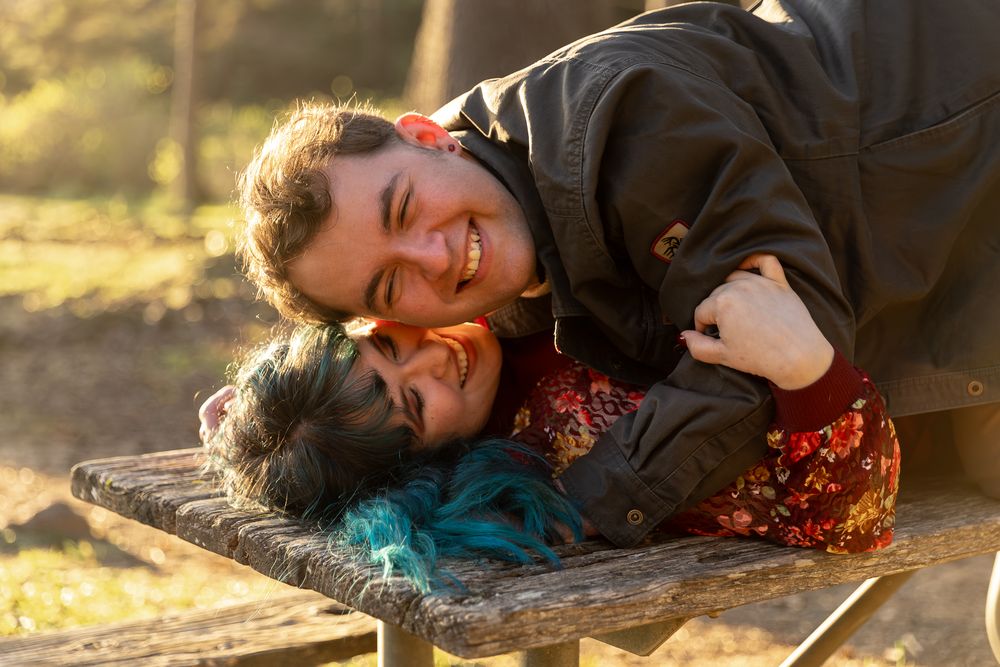 Two young people laughing and cuddling on a wooden picnic table in a sunny autumn park setting.