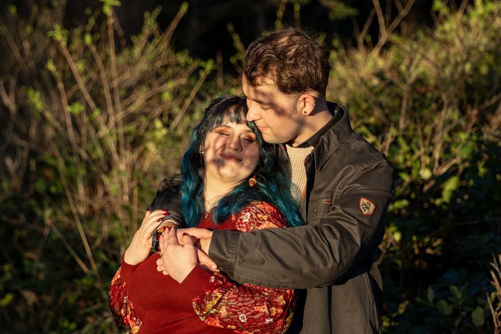 A couple embraces tenderly outdoors, woman with blue hair in red floral dress, man in gray jacket, surrounded by green foliage.