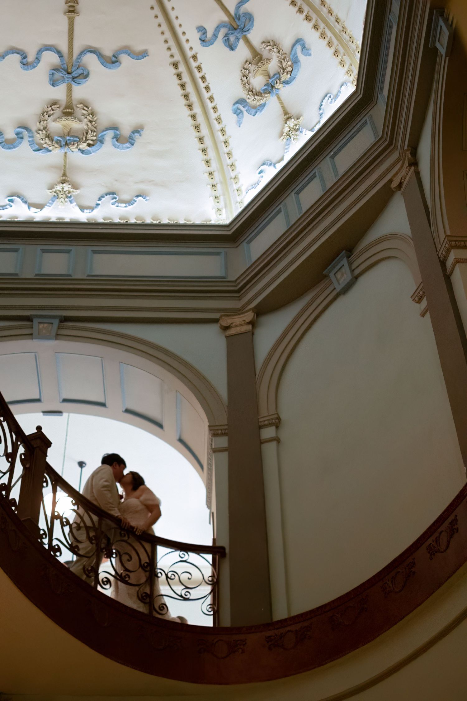 Couple embracing on ornate curved staircase beneath elegant decorative ceiling with blue floral motifs in historic building.