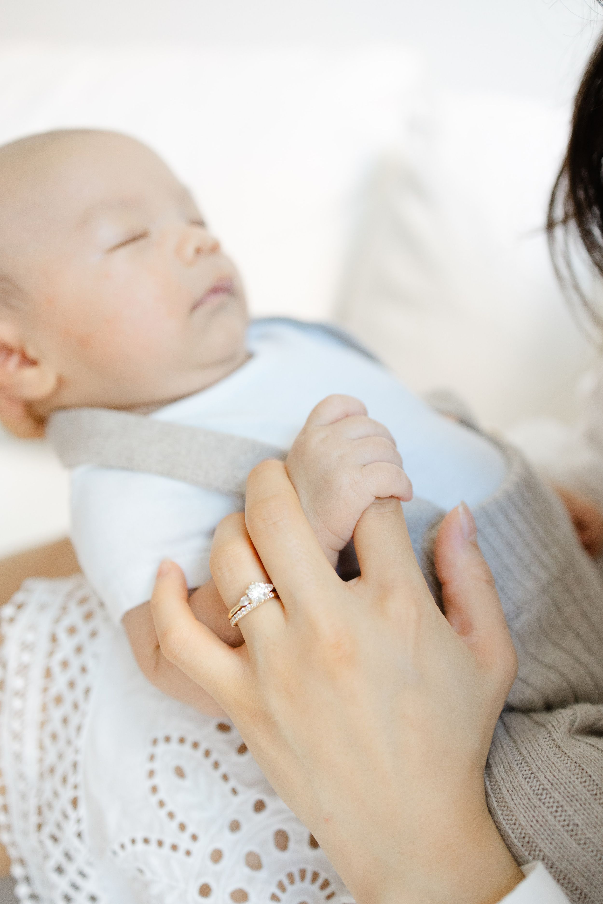 A mother tenderly holds her sleeping baby's hand, wearing a gold ring, while the infant rests in a light blue outfit.