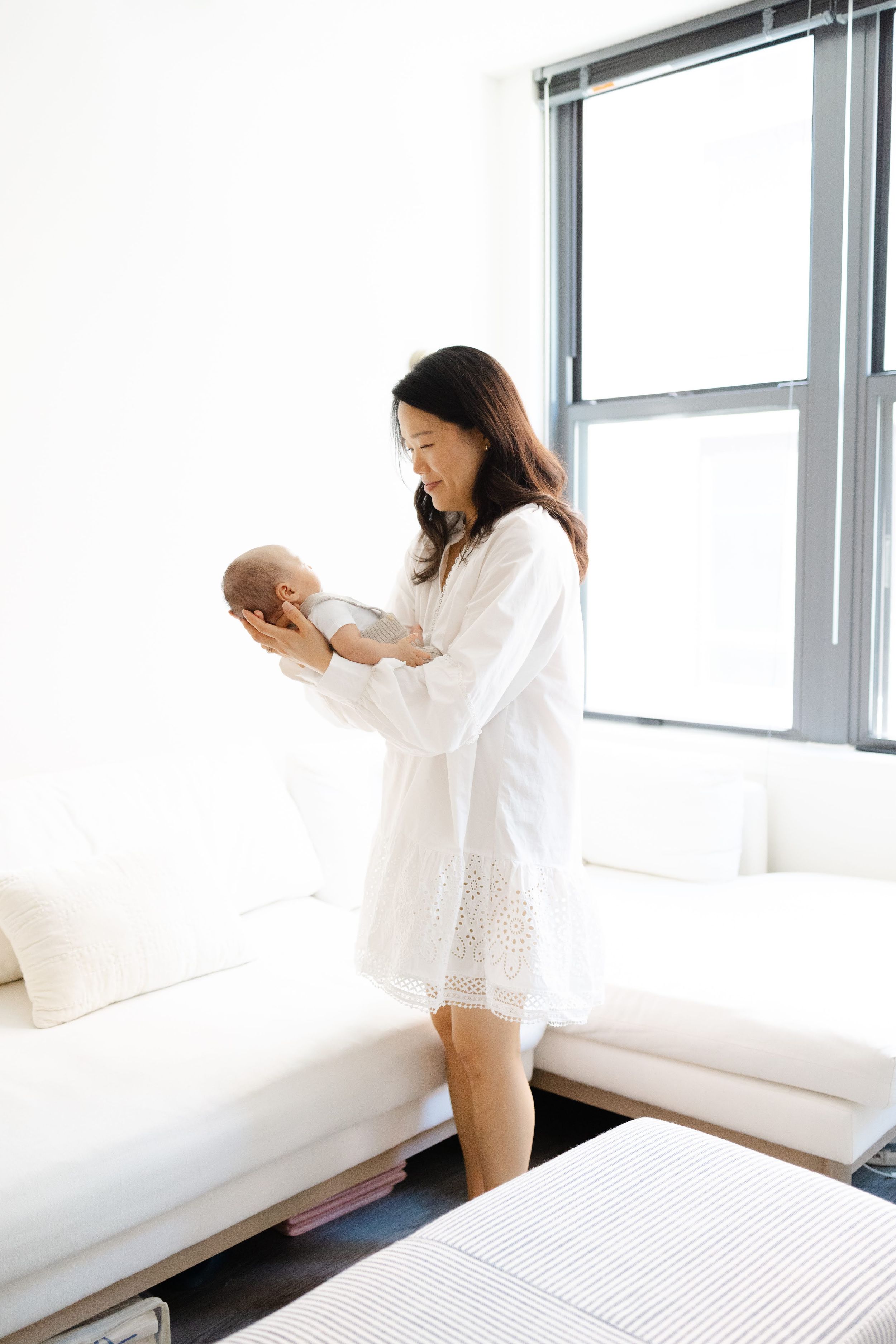 A woman in a white dress holds her newborn baby near a bright window in a modern living room.