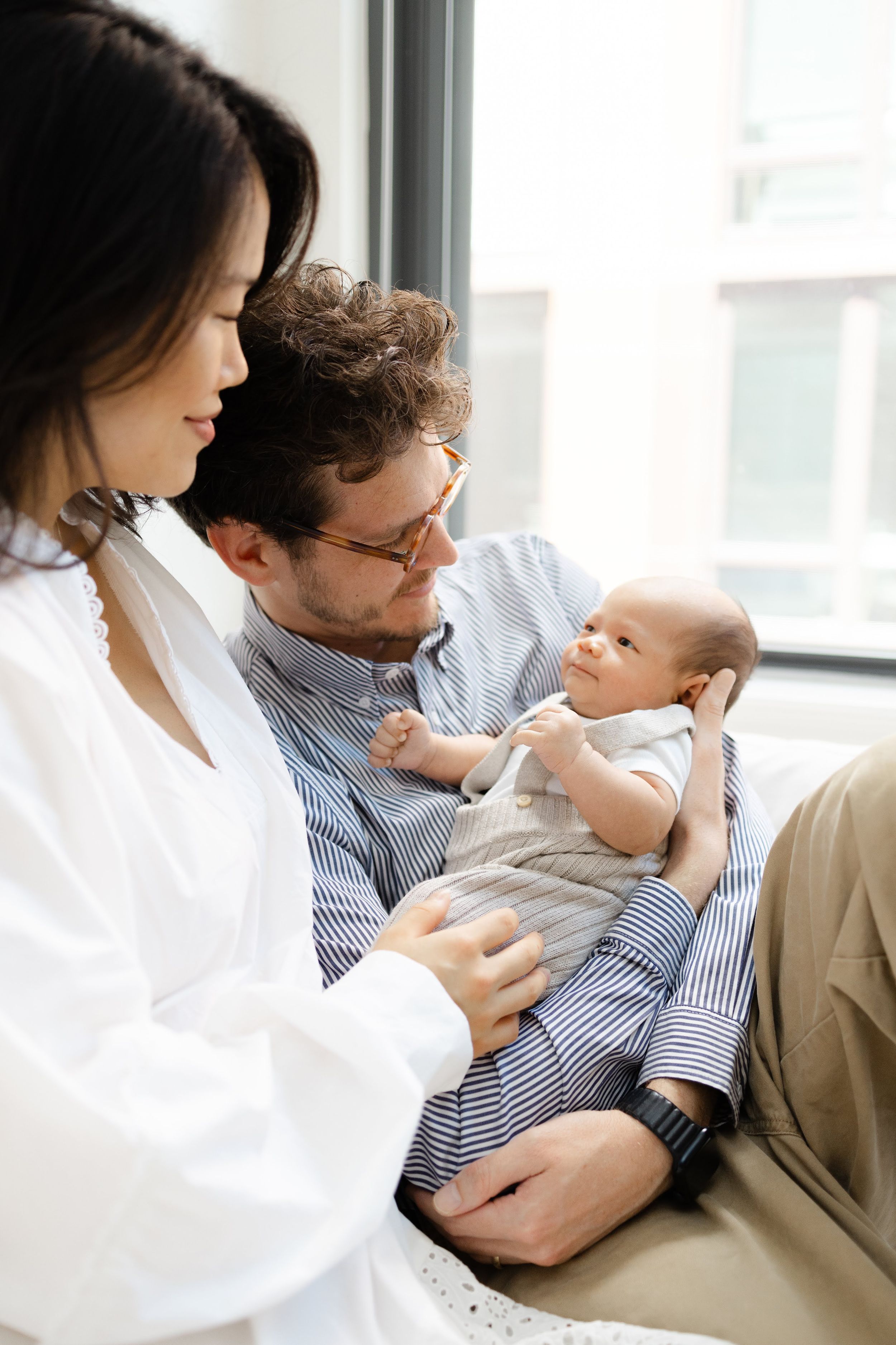 A young family with a newborn baby, mother in white blouse and father in striped shirt sitting by a window.