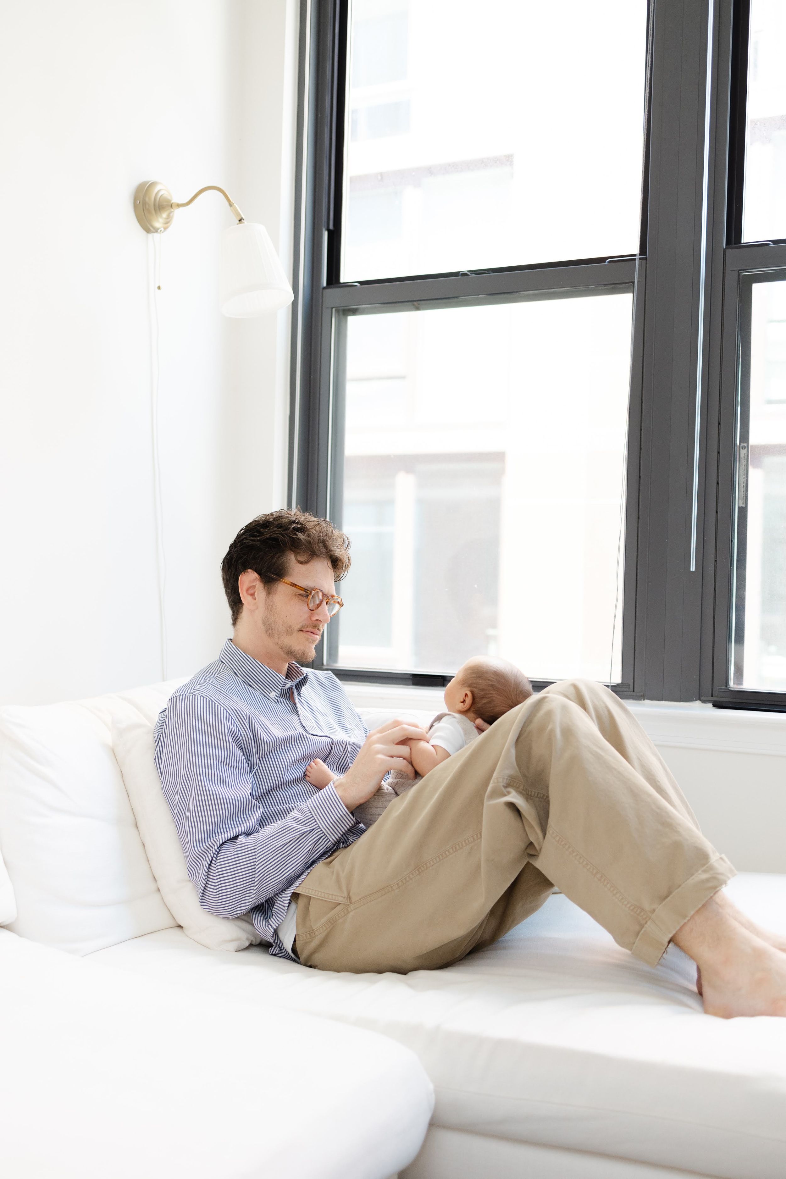 A man in glasses sits on a white couch cradling a newborn baby near large windows in a bright modern room.