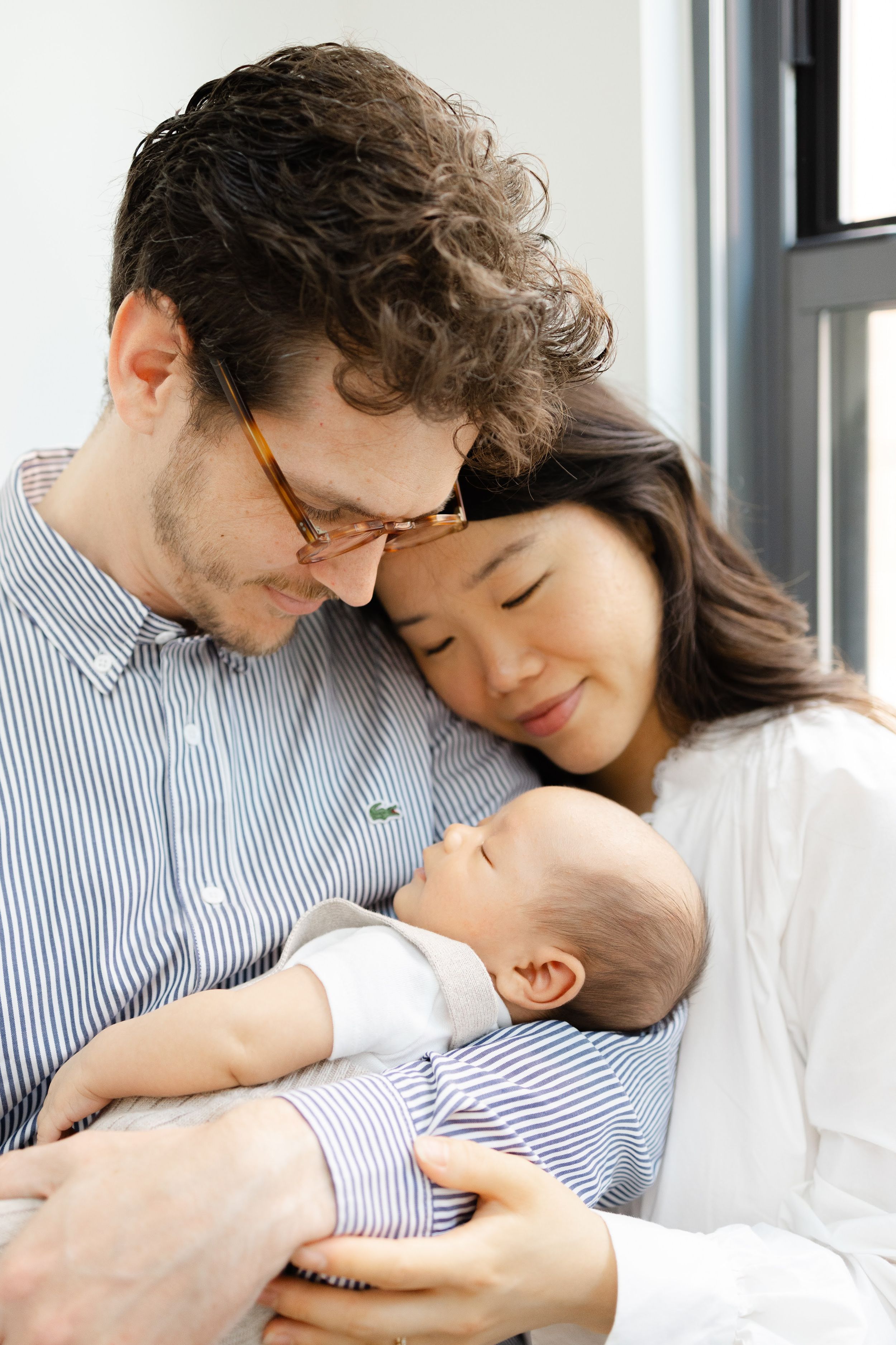 A couple tenderly gazes at their newborn baby, with the man wearing glasses and the woman leaning her head on his shoulder.