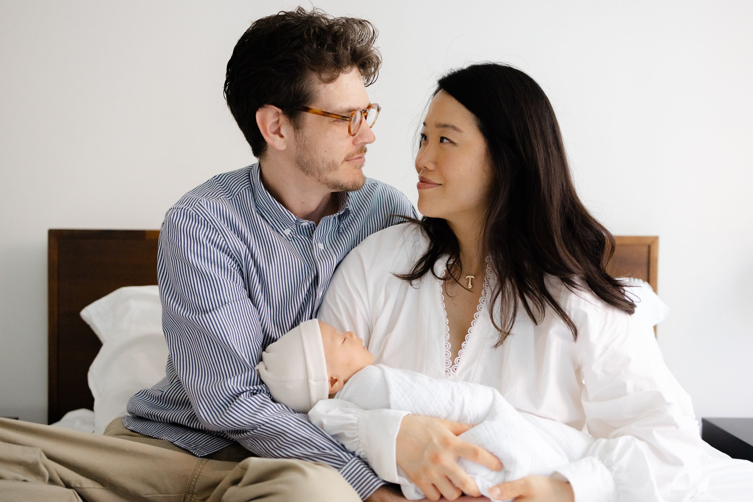 A couple sits on a bed smiling at each other while tenderly holding their newborn baby.