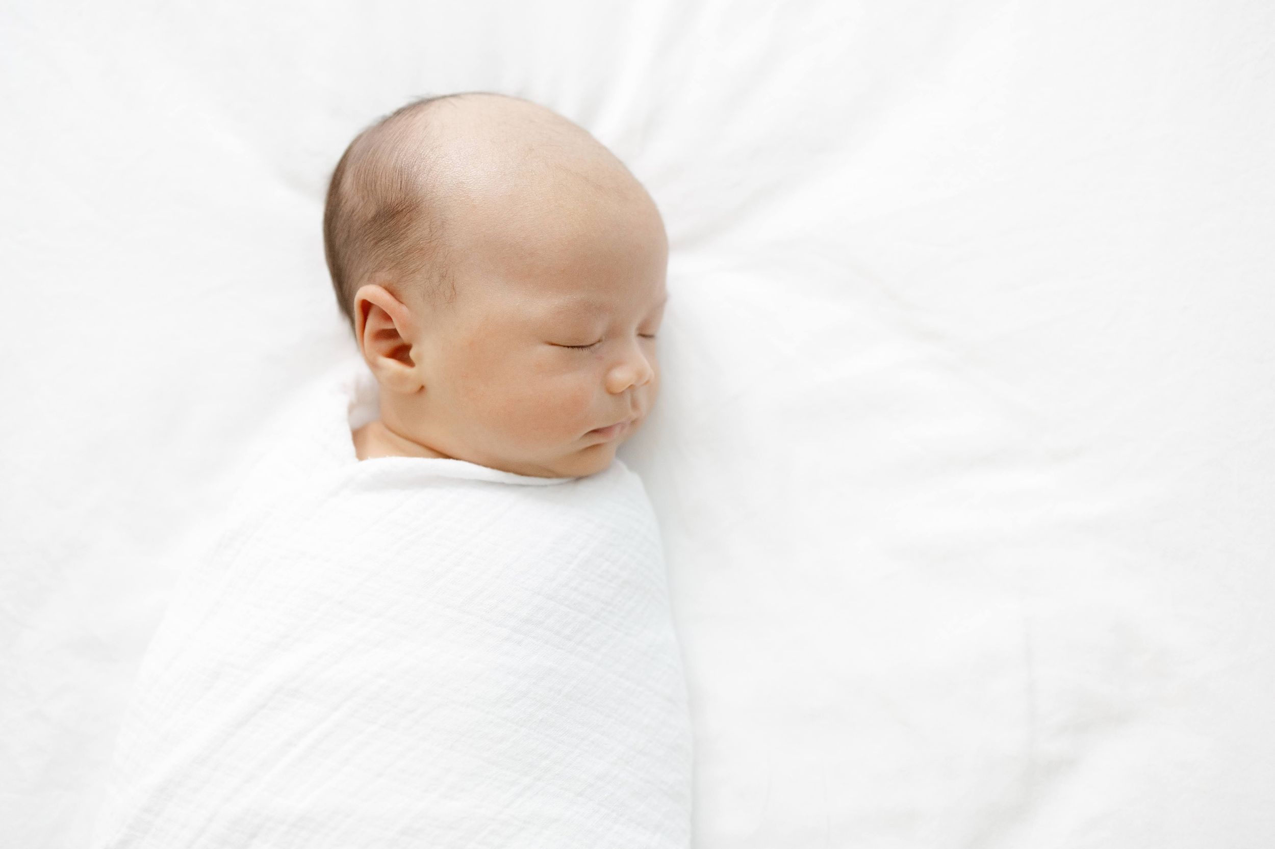 A peaceful newborn baby swaddled in a white muslin wrap, sleeping soundly on a soft white blanket.