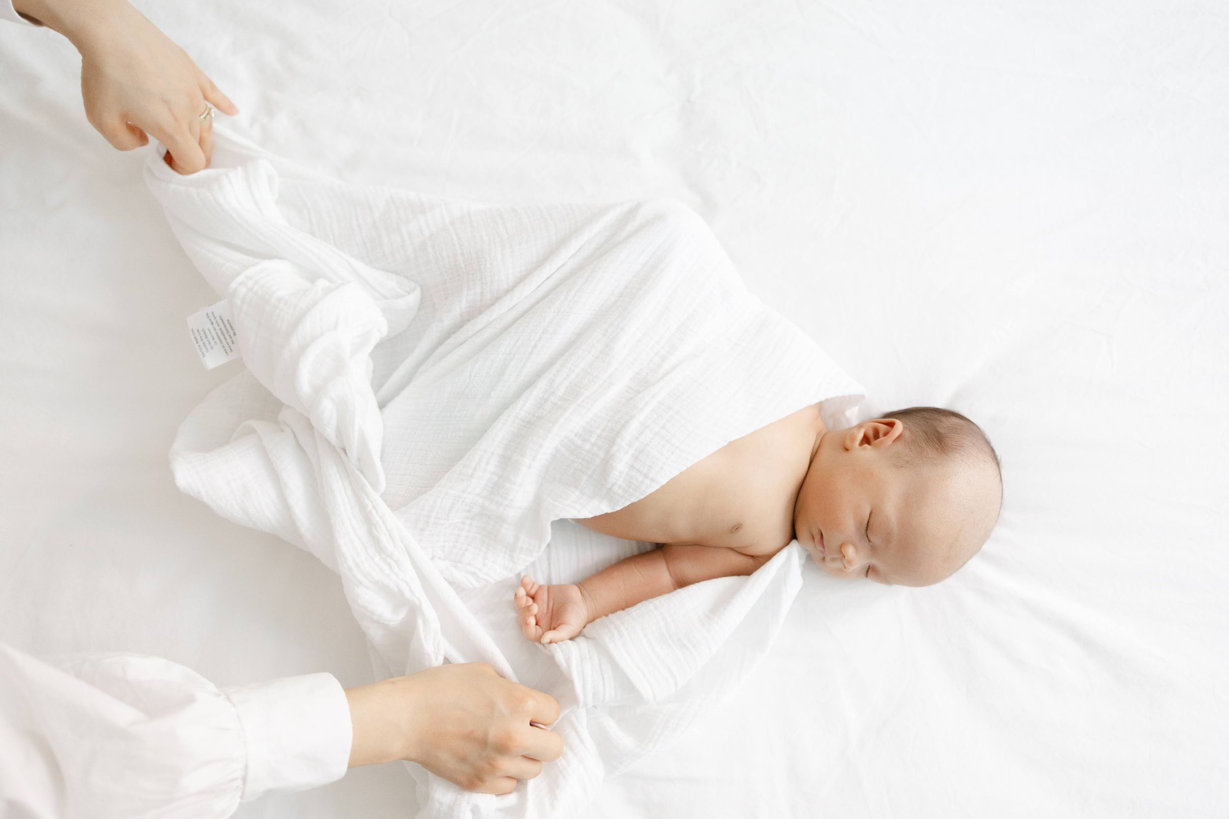 Newborn baby sleeping peacefully on white bedding while caregiver gently wraps them in a soft white cloth.