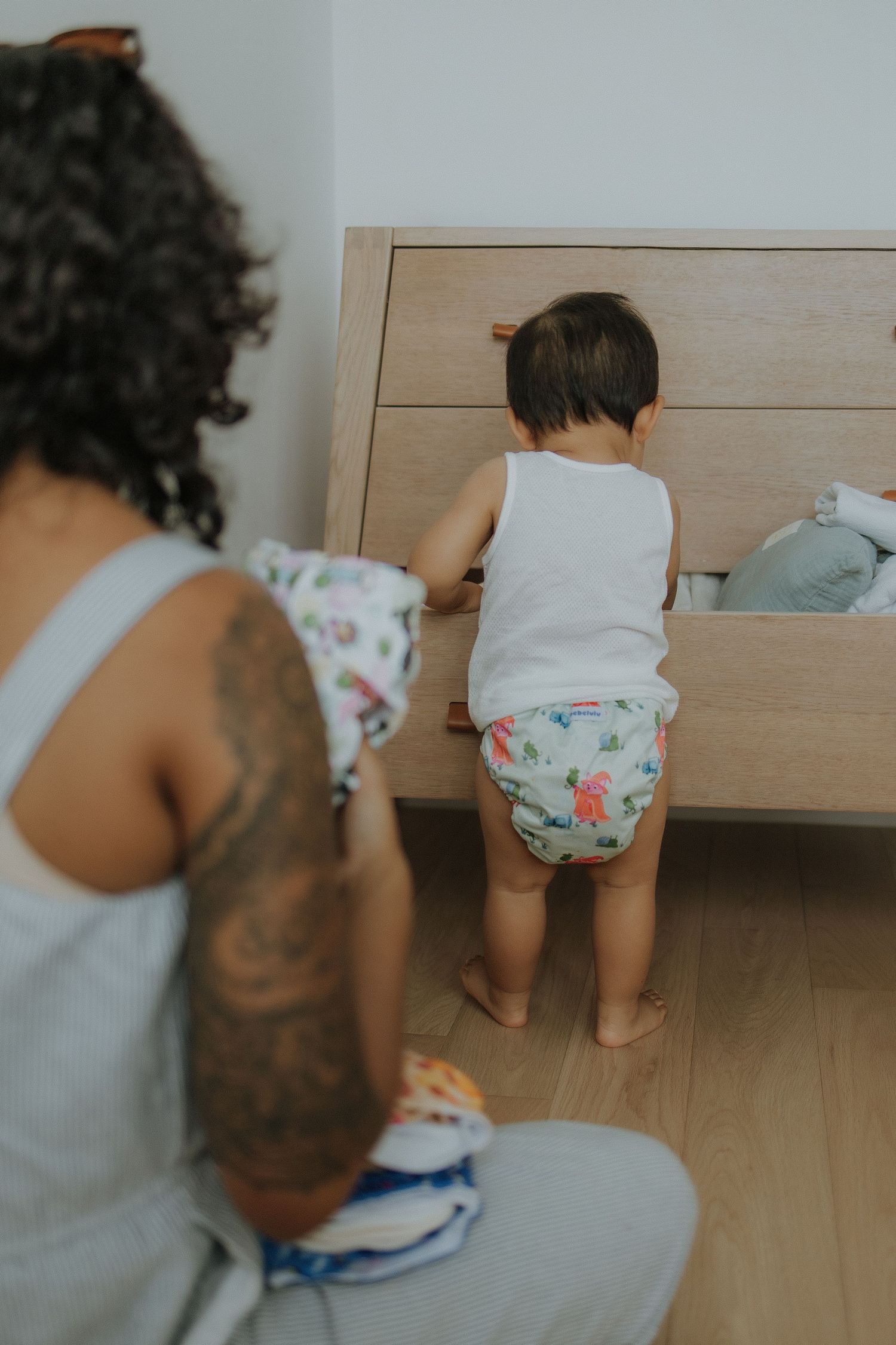 Toddler in diaper and tank top exploring wooden dresser drawer while tattooed parent watches nearby.