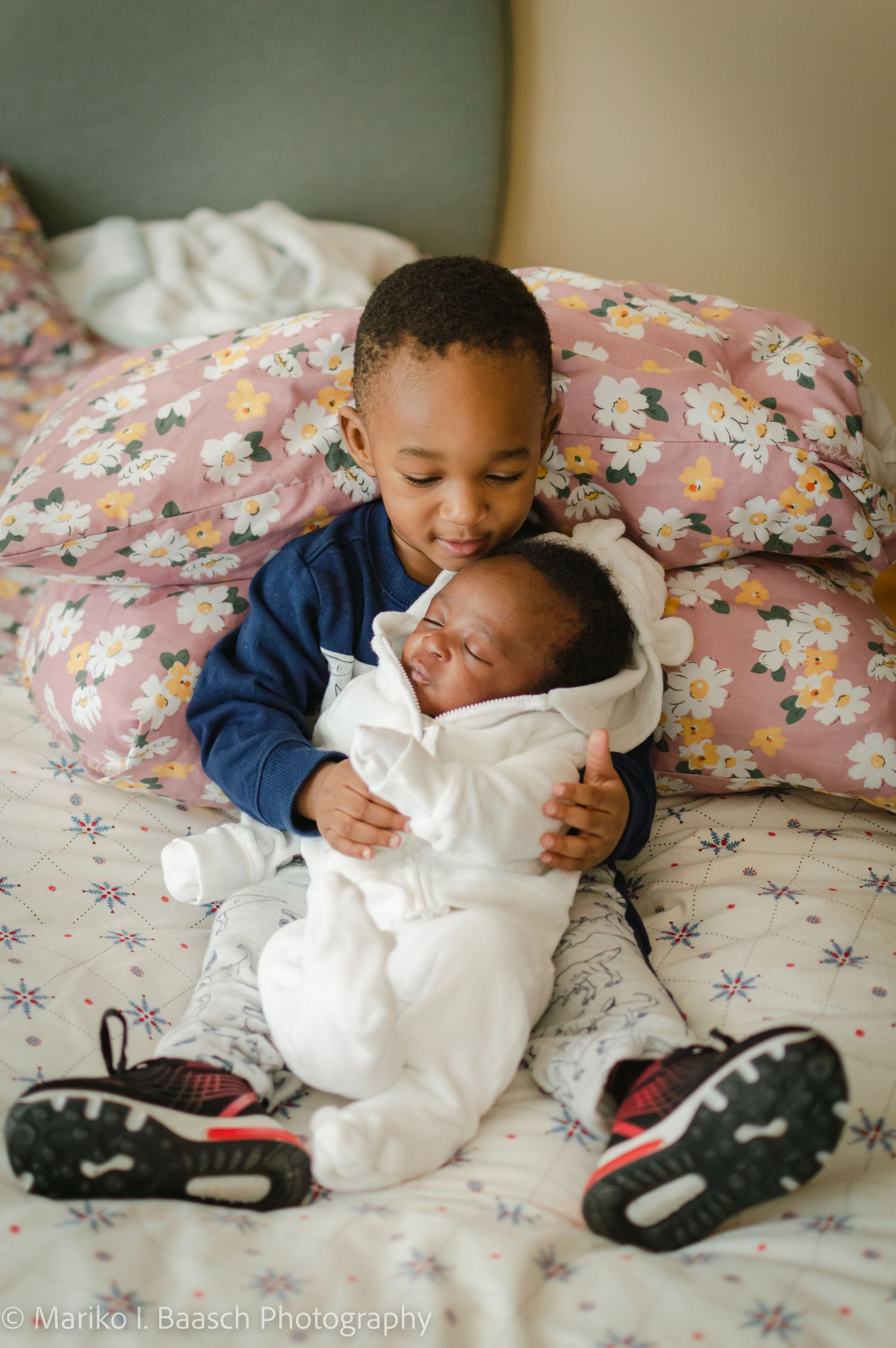 A young boy tenderly holds his sleeping newborn sibling on a floral bedspread in a sweet sibling portrait.