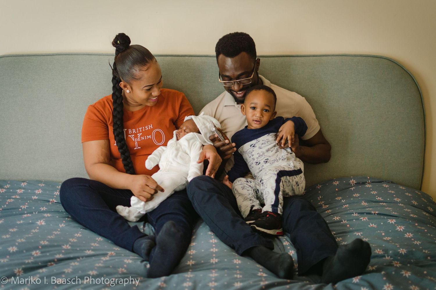 A happy family of four sits on a bed, parents holding their newborn and toddler during a newborn photo session.