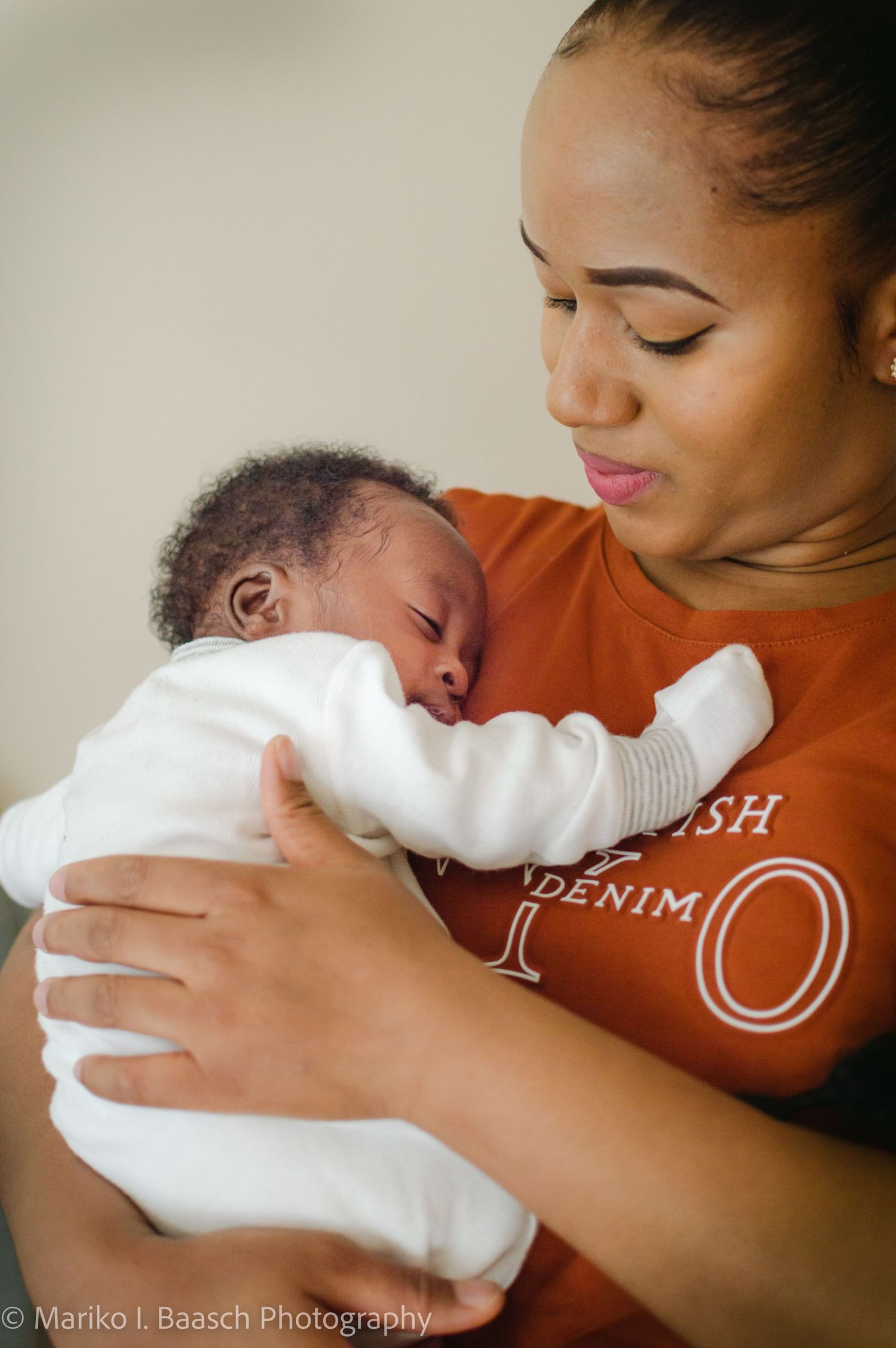 A mother in an orange shirt gently cradles her sleeping newborn baby close to her chest in a tender portrait.