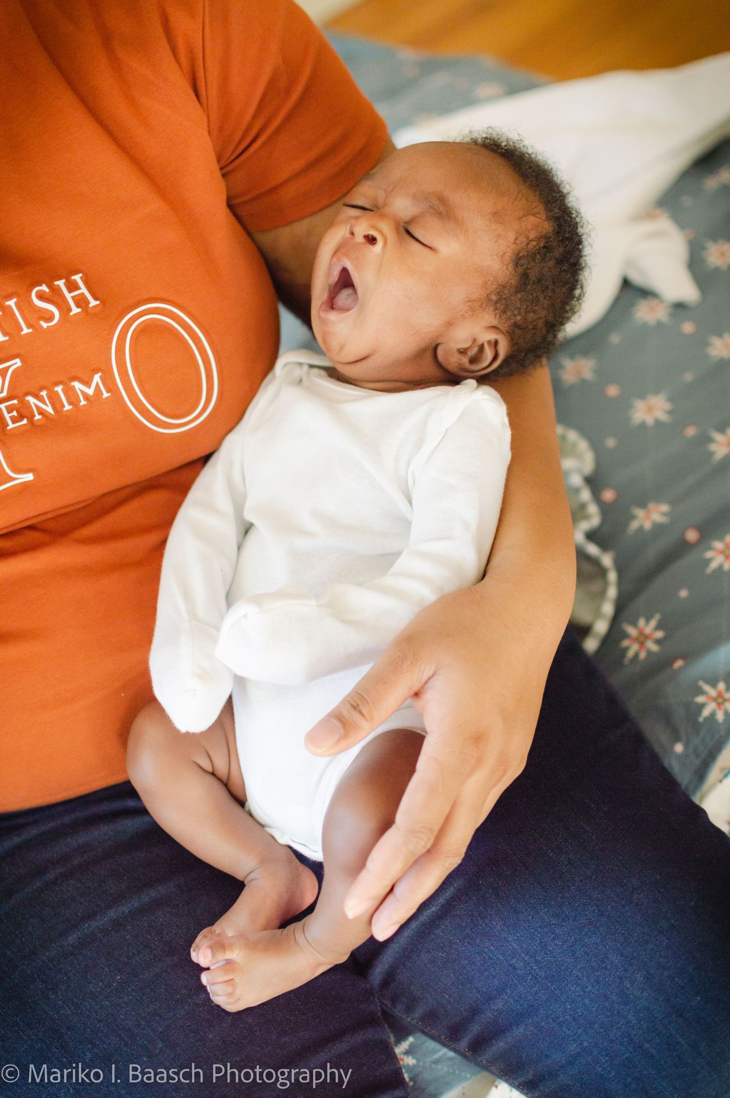 A newborn baby yawns widely while being held by their mother in an orange shirt during a home photo session.