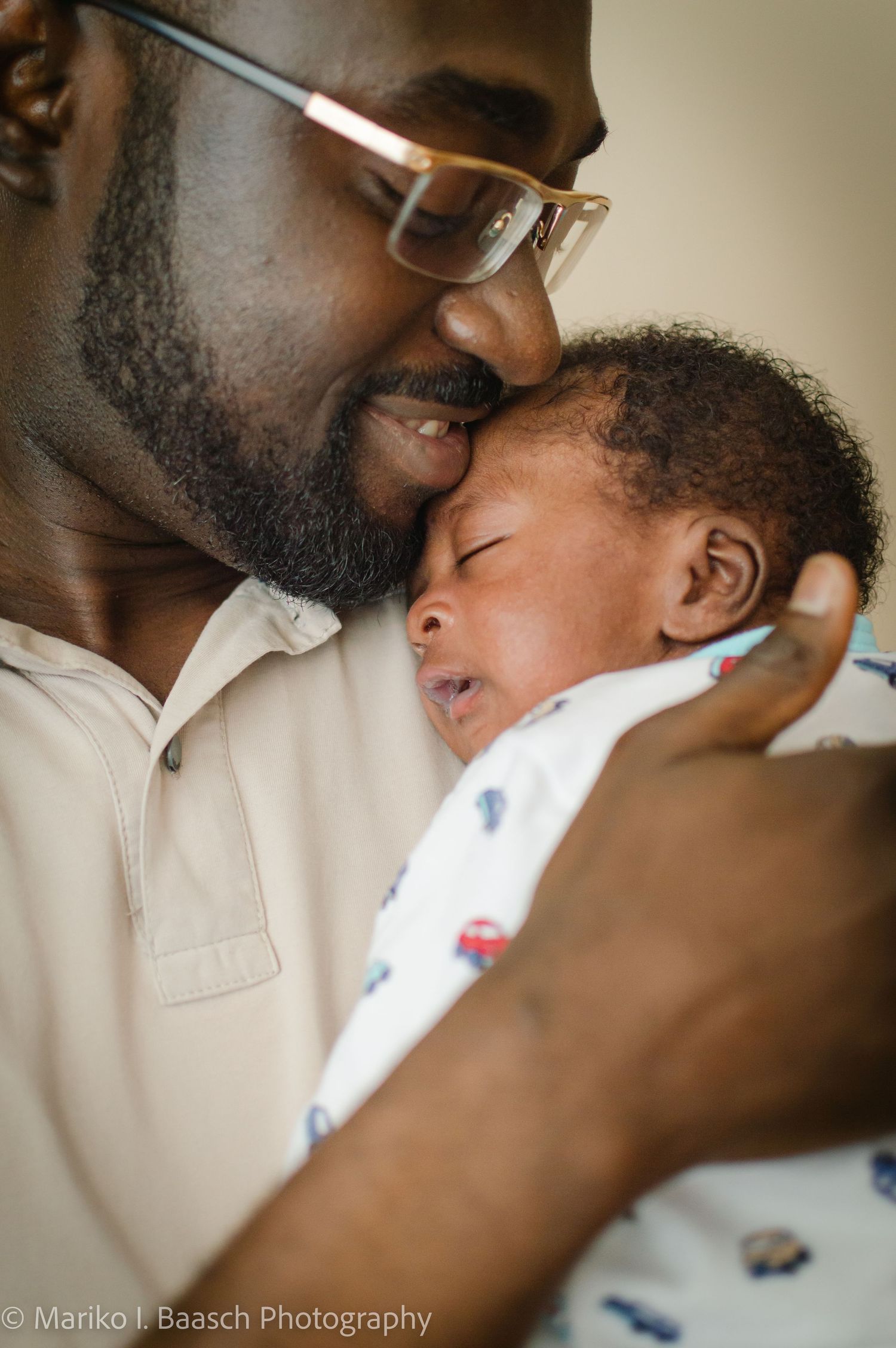 A father wearing glasses shares a tender cheek-to-cheek moment with his crying newborn in a close-up portrait.