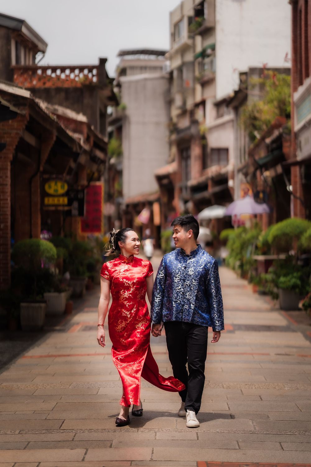 Couple in traditional Chinese attire walking through a historic street, woman in red qipao and man in blue tang suit.