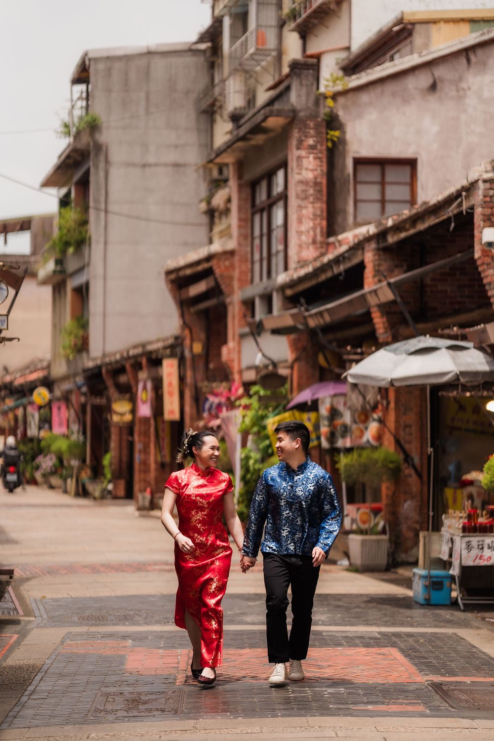Couple in traditional Chinese attire walking hand in hand through a historic Asian street market.