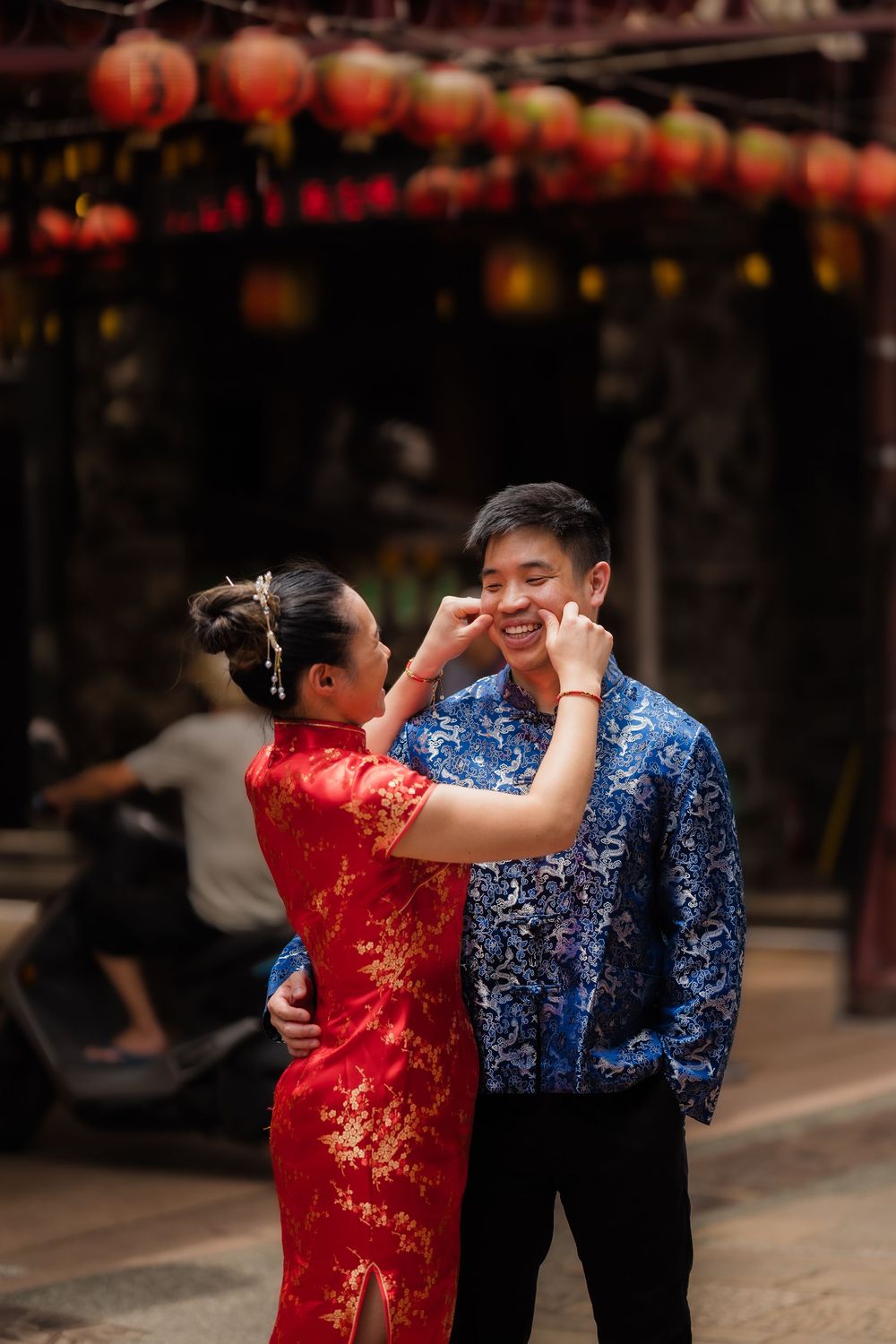 Couple in traditional Chinese attire embrace under red lanterns, woman in red qipao touches mans smiling face.