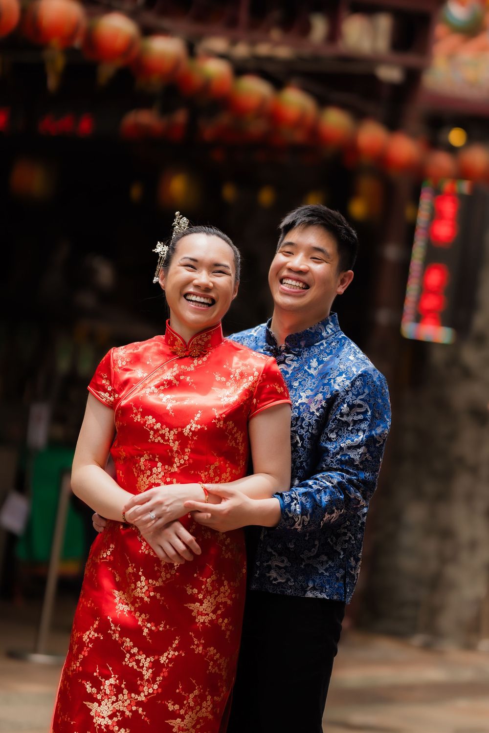 Happy Asian couple in traditional Chinese attire embracing, woman in red qipao, man in blue, with red lanterns behind.