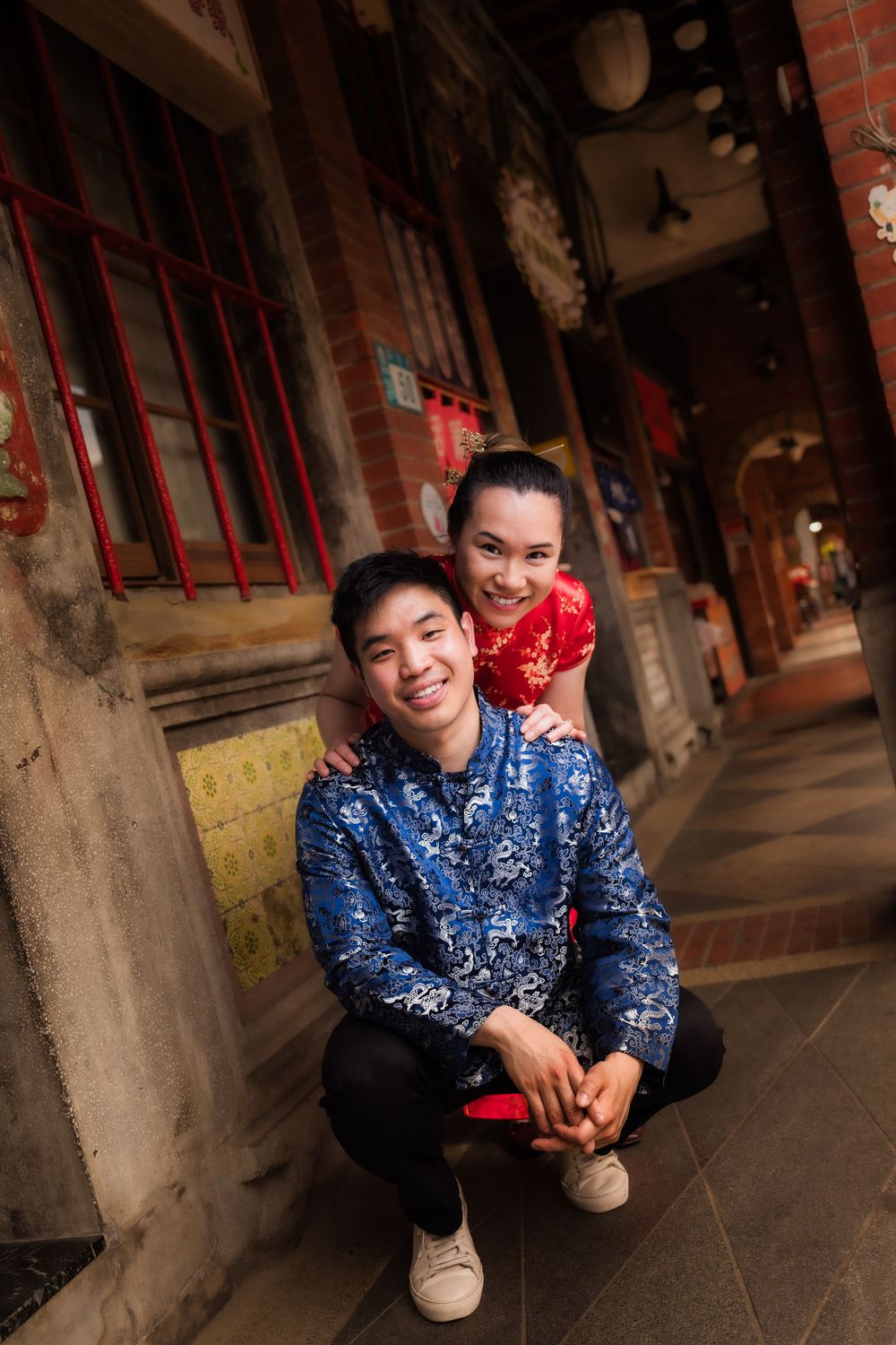 A young Asian couple in traditional Chinese attire pose together in a historic alleyway, smiling warmly at the camera.