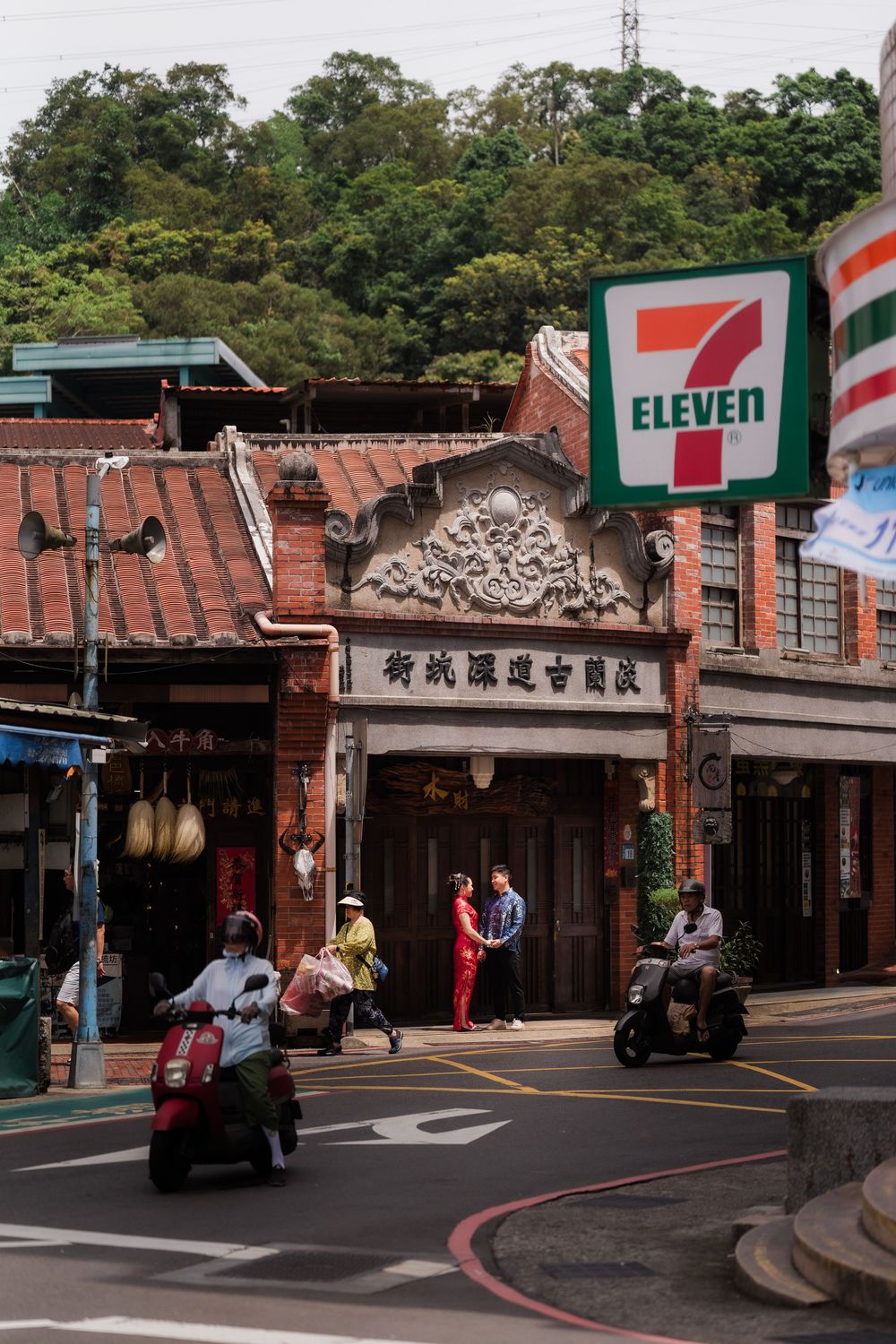 Historic Taiwanese street scene with 7-Eleven sign, traditional architecture, and motorbikes passing by.