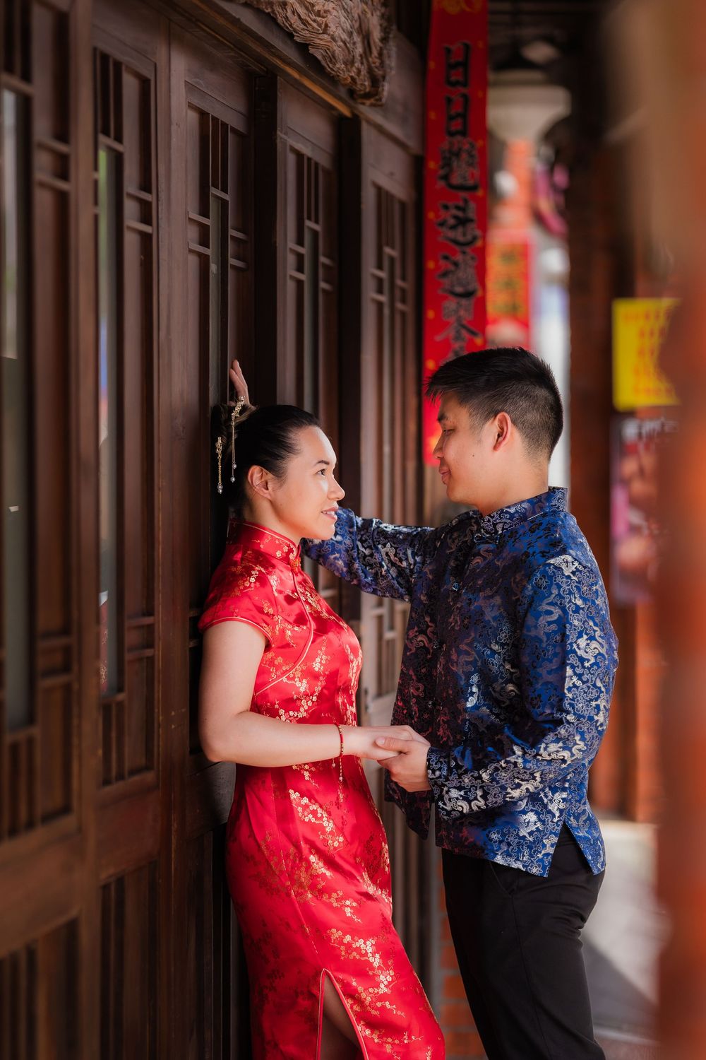 Couple in traditional Chinese attire, woman in red qipao and man in blue floral top, posing in a wooden corridor.