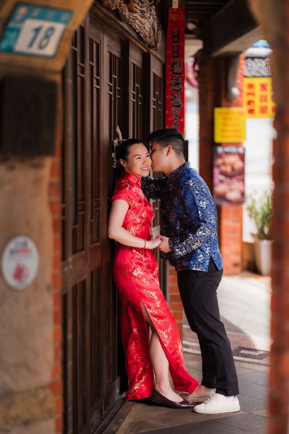 Couple in traditional Chinese attire, woman in red qipao, man in blue, posing romantically by wooden doors in Chinatown.