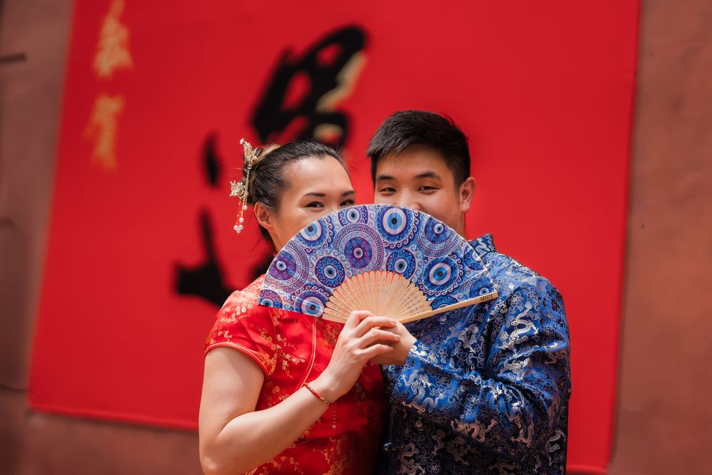 Couple in traditional Chinese attire posing with a decorative blue fan against a red background.