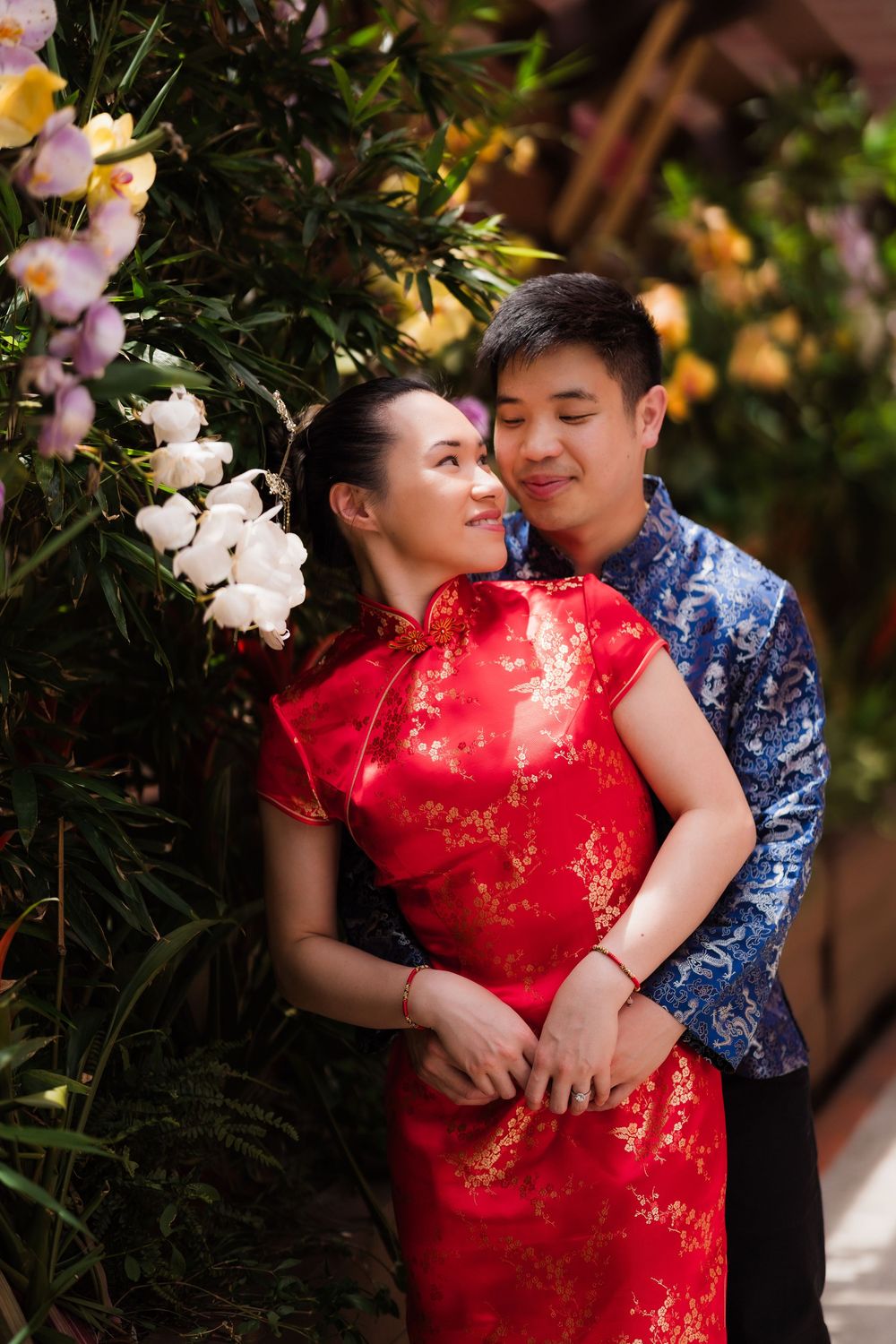 Romantic couple in traditional Chinese red and blue outfits posing near blooming flowers.