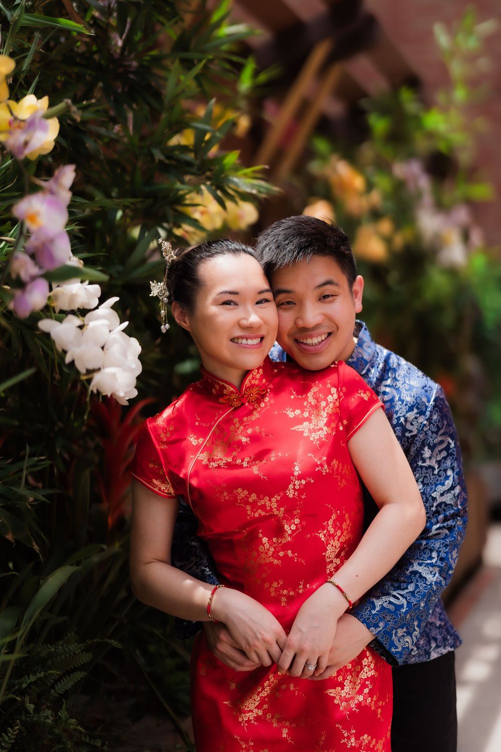 Couple in traditional Chinese attire, red qipao and blue top, smiling among blooming flowers.