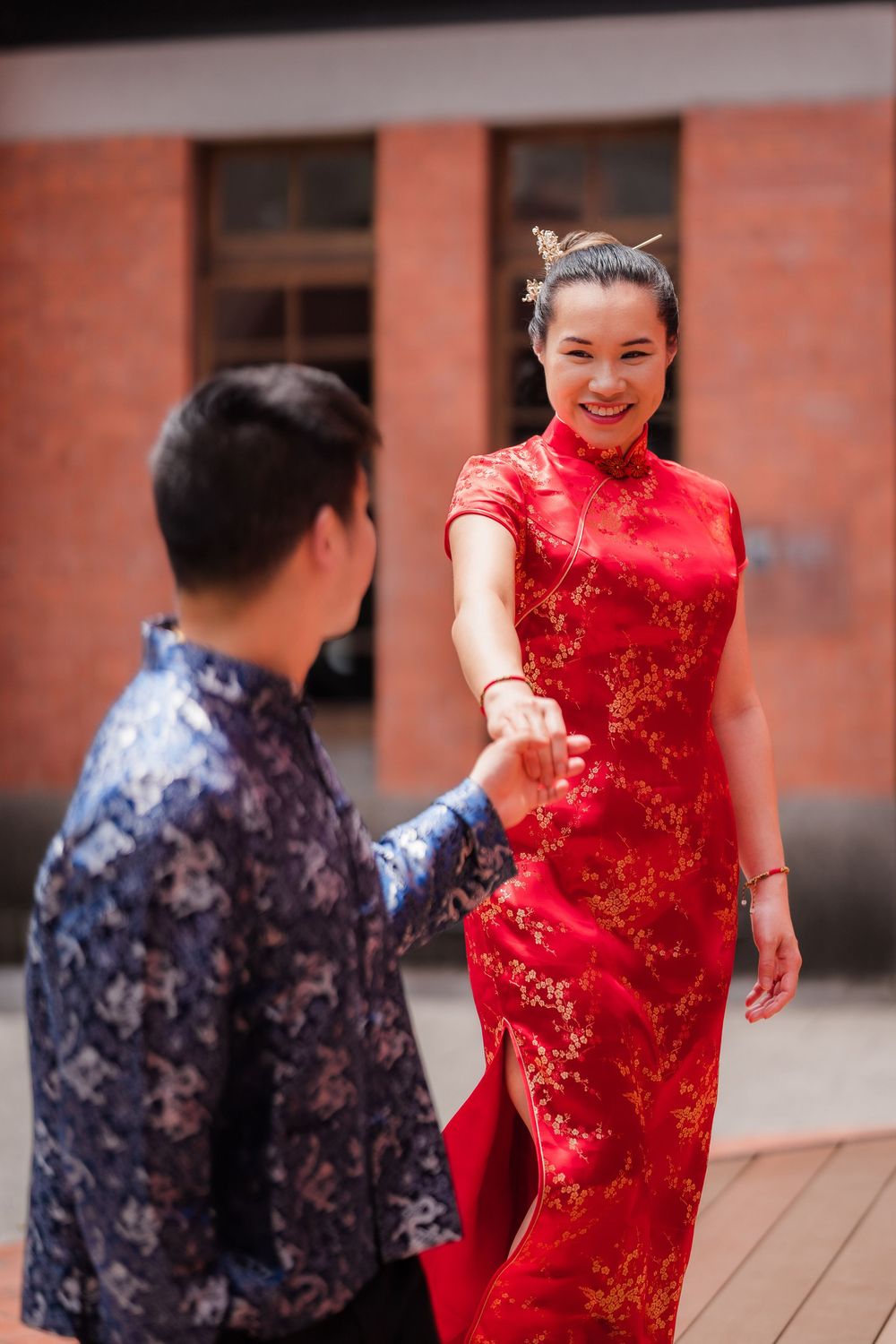 Smiling woman in red qipao holding hands with man in blue Chinese top outdoors near brick building.