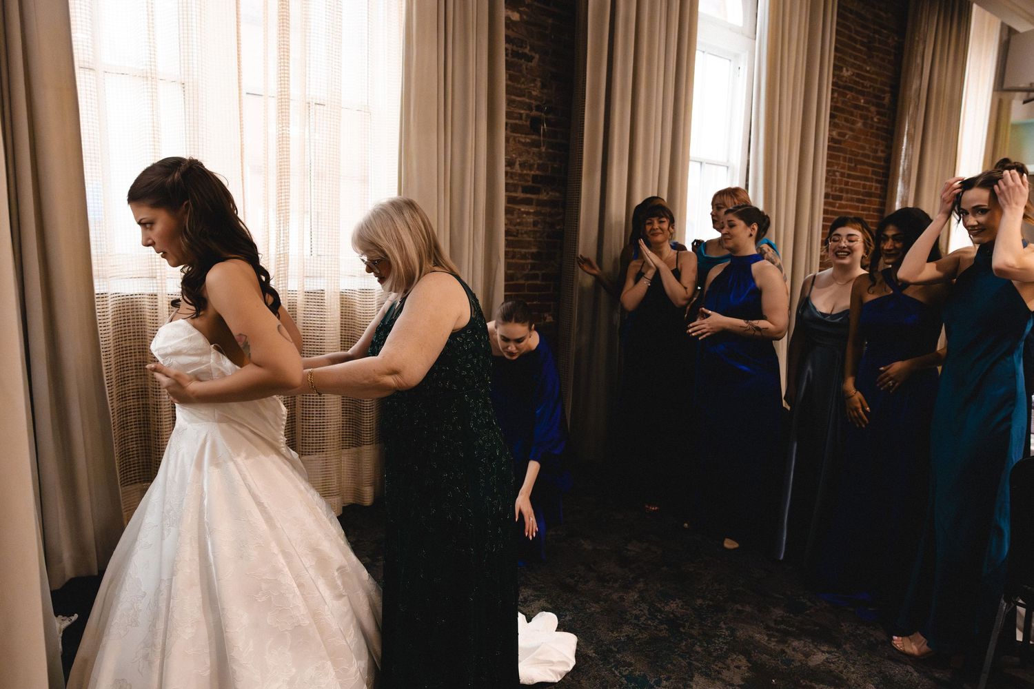 Bride getting dressed with bridesmaids in blue gowns cheering in elegant venue with brick walls and large windows.