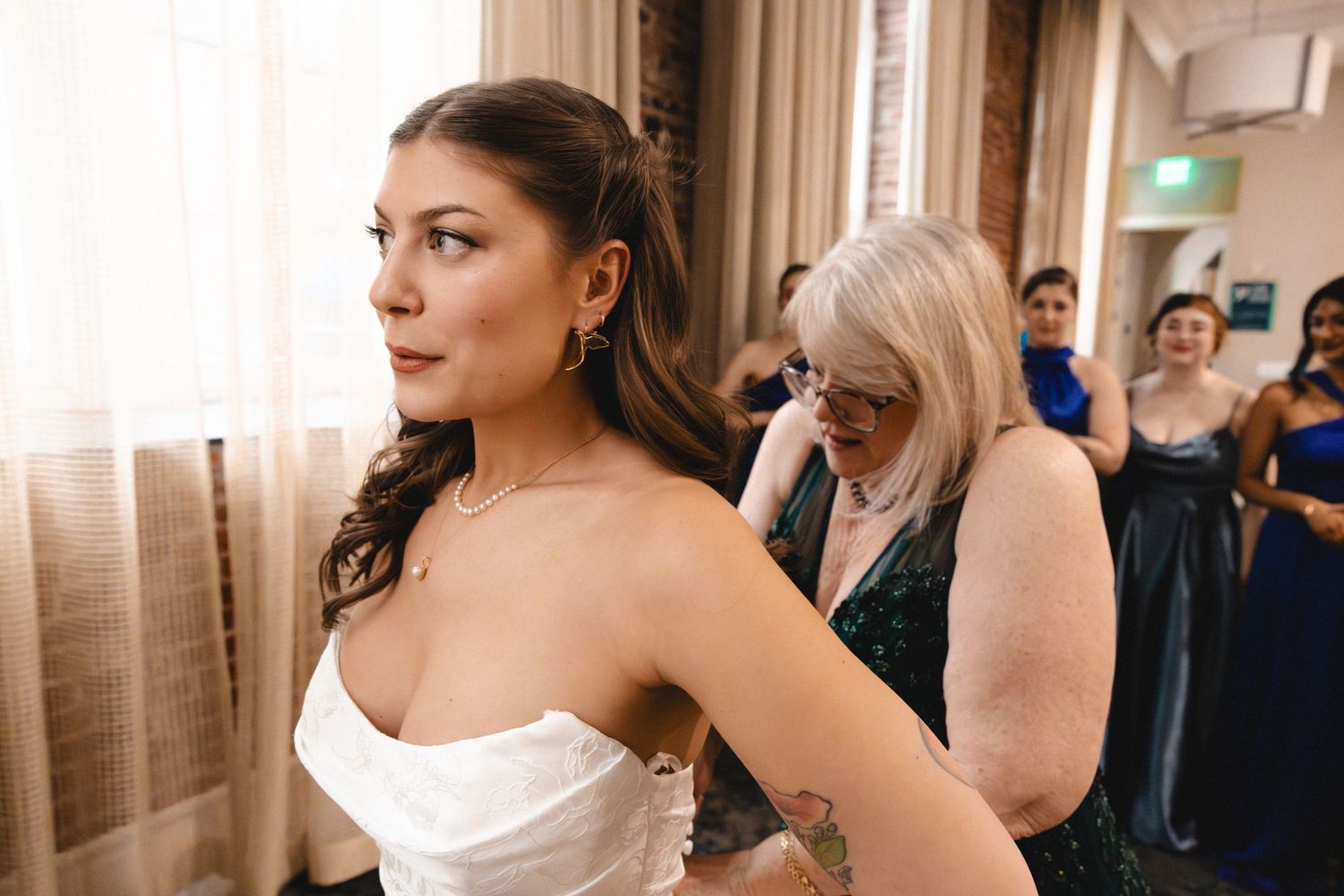 Bride in white strapless gown being helped by woman in green dress, with wedding guests in background.
