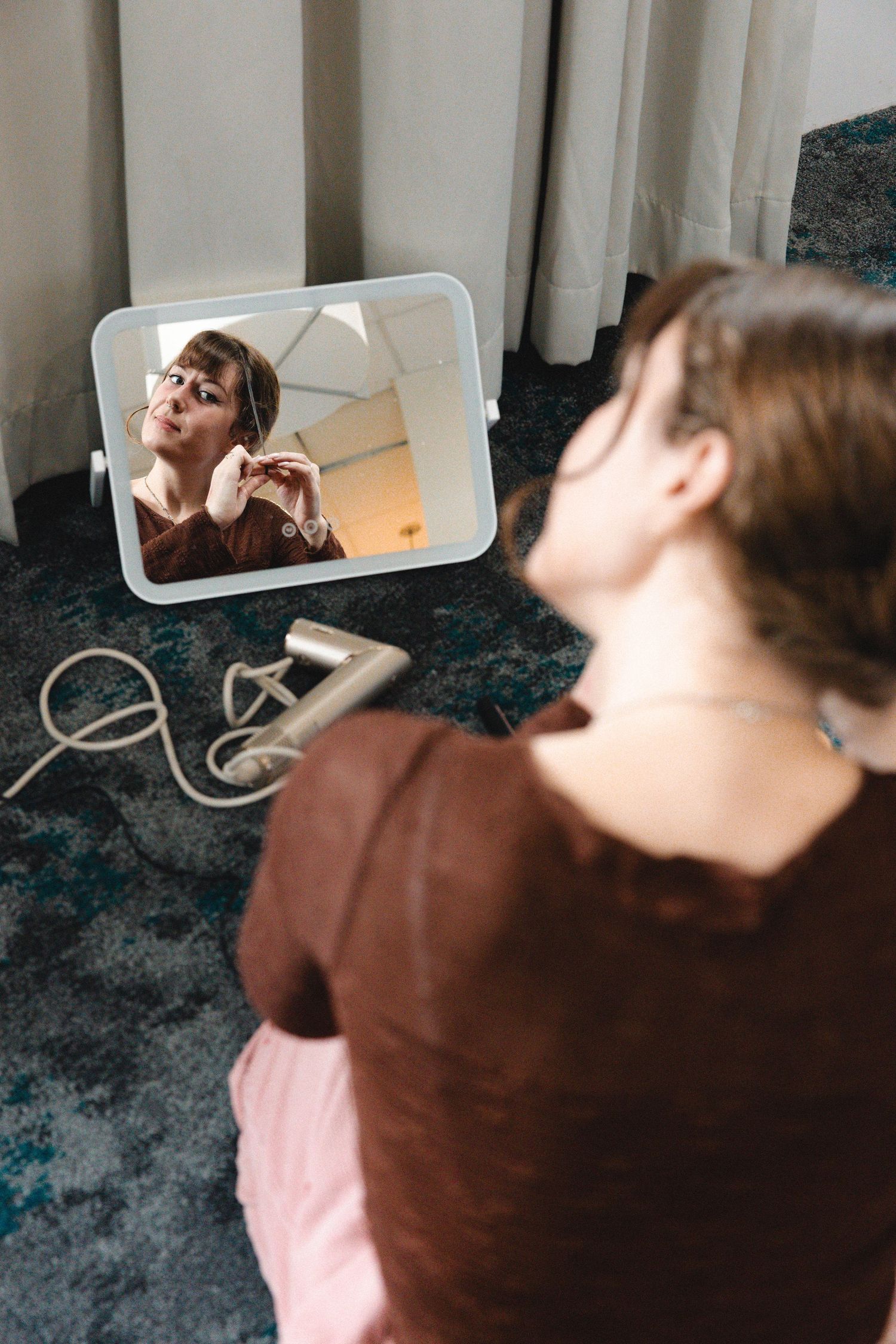 Woman sitting on floor applying makeup while looking at her reflection in a small mirror on carpet.