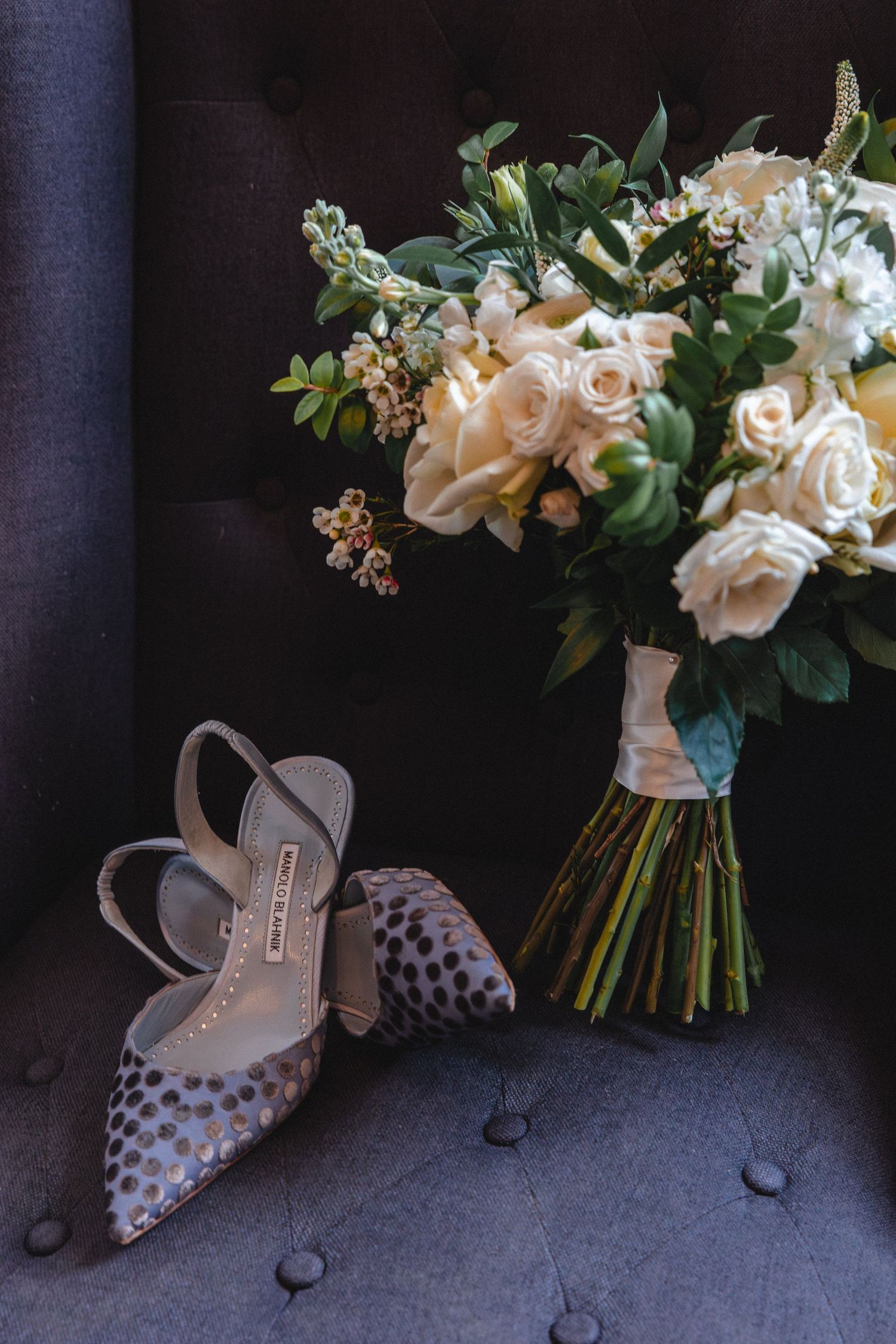 Elegant bridal shoes with gold polka dots beside a lush white rose and greenery wedding bouquet on a gray velvet chair.