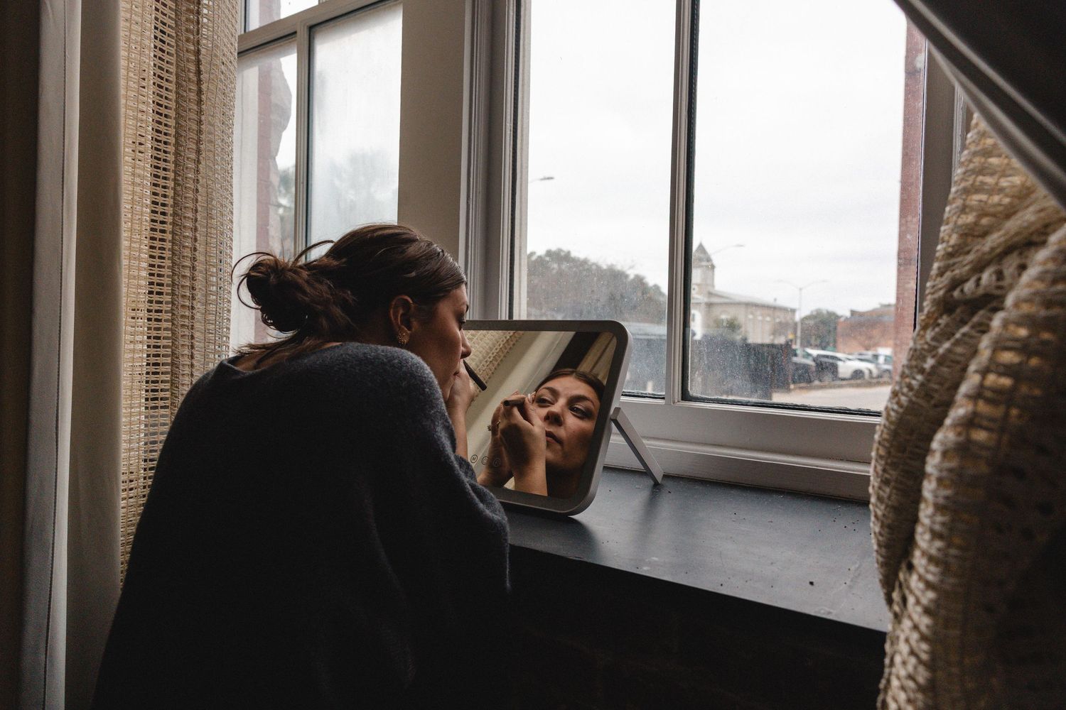 Woman applying makeup while looking into a small mirror on a desk by a window with a city view.