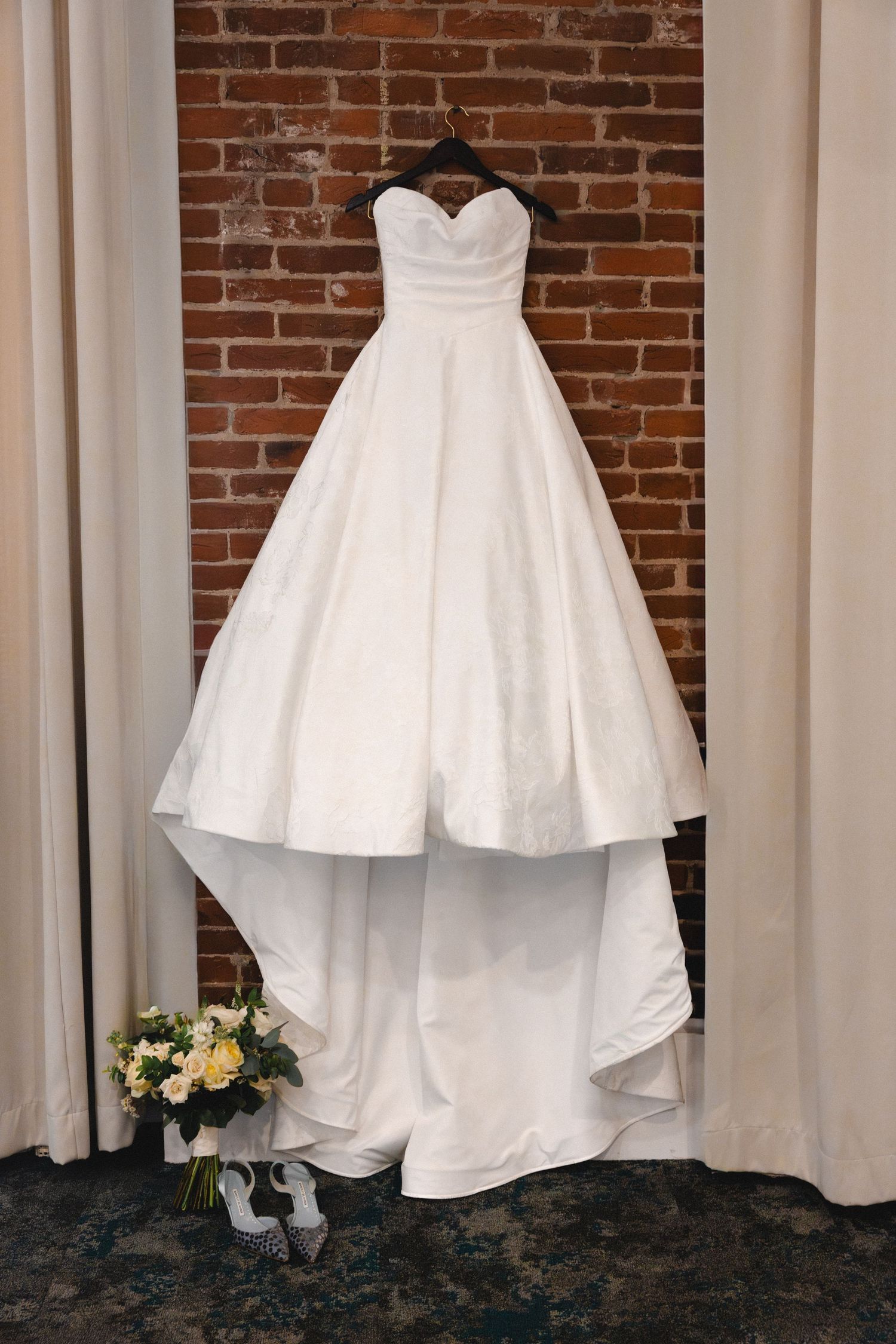 White strapless ballgown wedding dress hanging on brick wall with bridal bouquet and heels below.