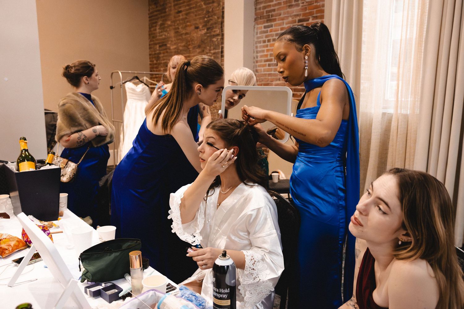 Bridesmaids in blue dresses help bride in white robe with hair and makeup in a bridal suite.