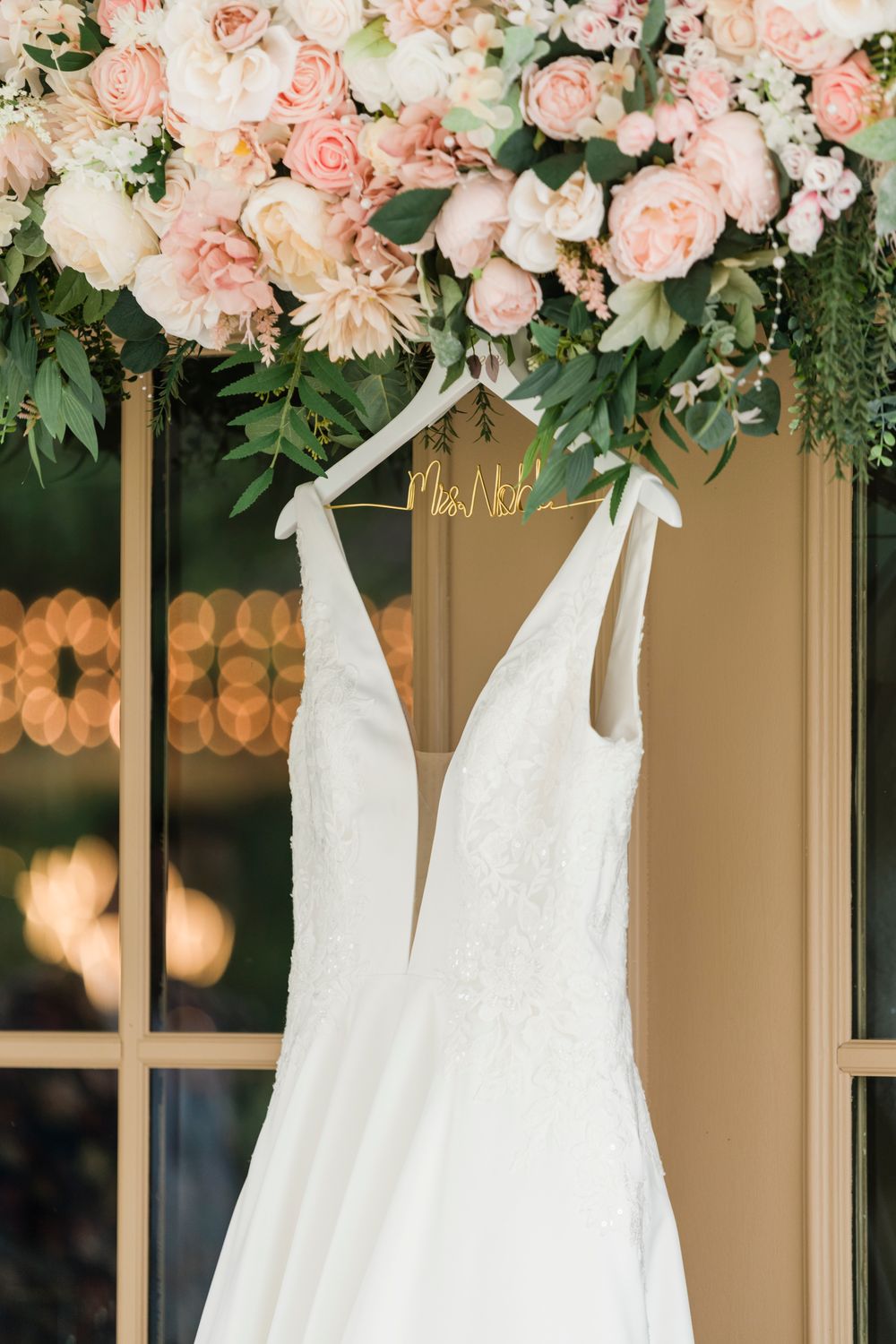 White lace wedding gown hanging on a personalized hanger beneath blush pink floral arrangement near elegant French doors.